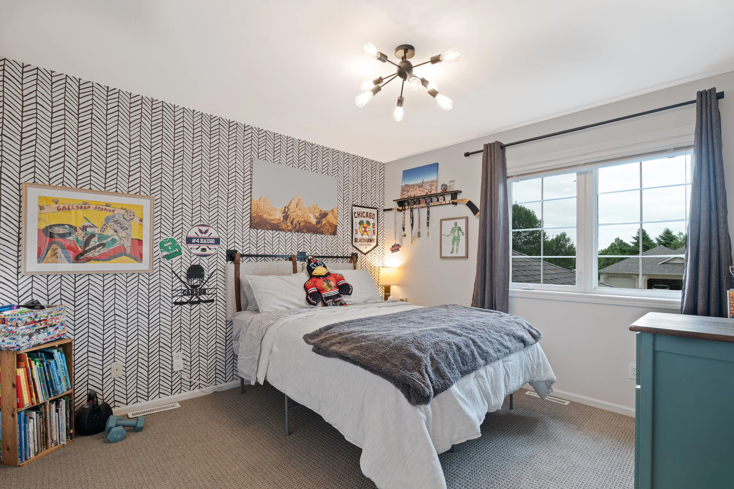 Bedroom with geometric accent wall, bed with white bedding and gray blanket, stuffed animal, artwork, window with views of trees and rooftops, and bookshelf.
