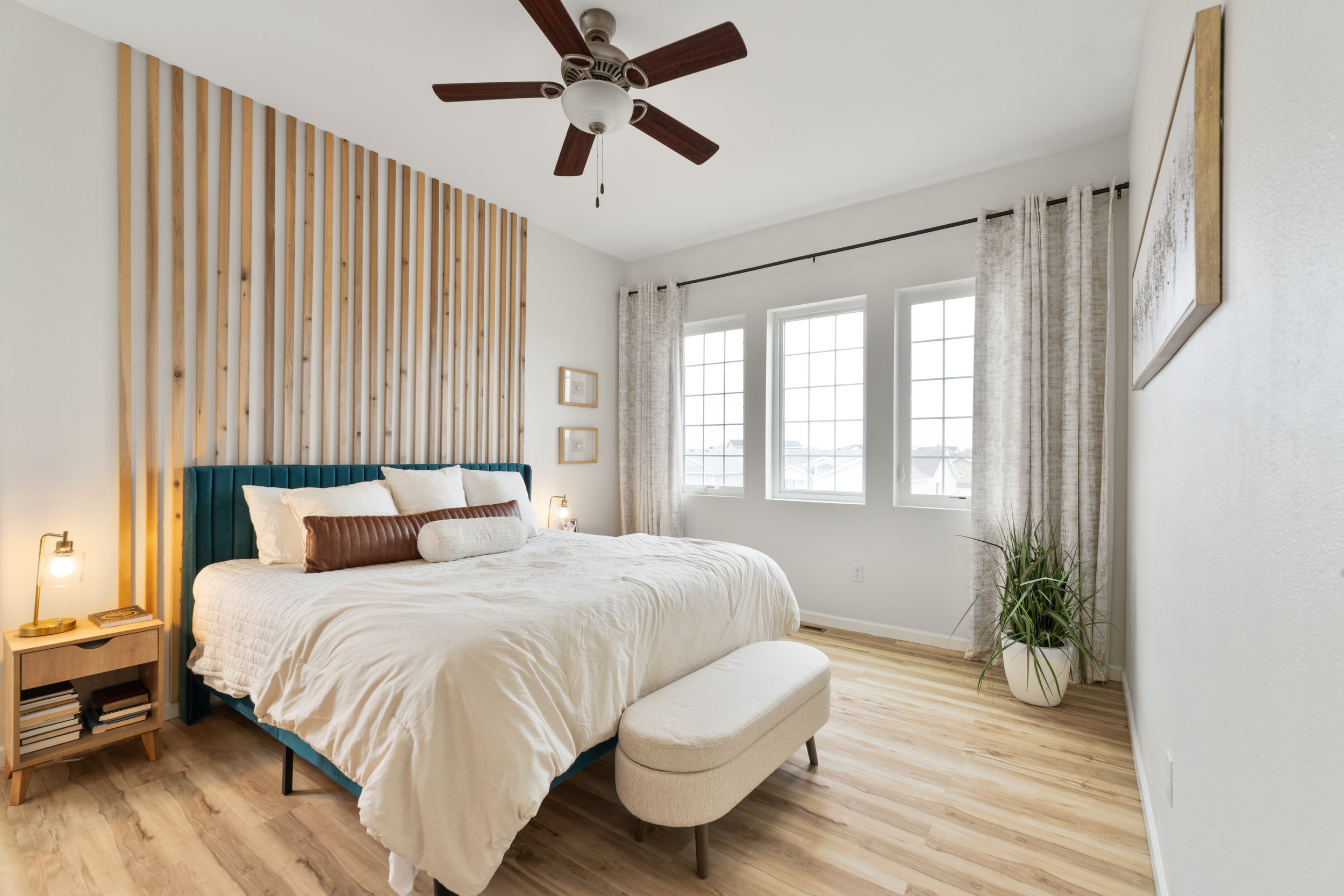 Bright bedroom with a large bed with white bedding, a teal upholstered headboard, and various pillows. A small wooden nightstand with books and a lamp is beside the bed. The room has three windows with white curtains, a ceiling fan with wooden blades