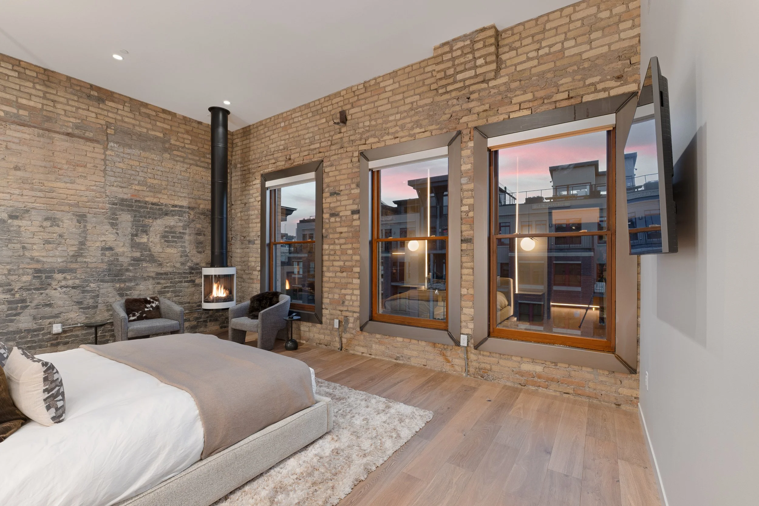 Bedroom with exposed brick wall, three windows showing an outside view, a fireplace, a bed with pillows, two chairs, a shaggy rug, and a mounted TV.