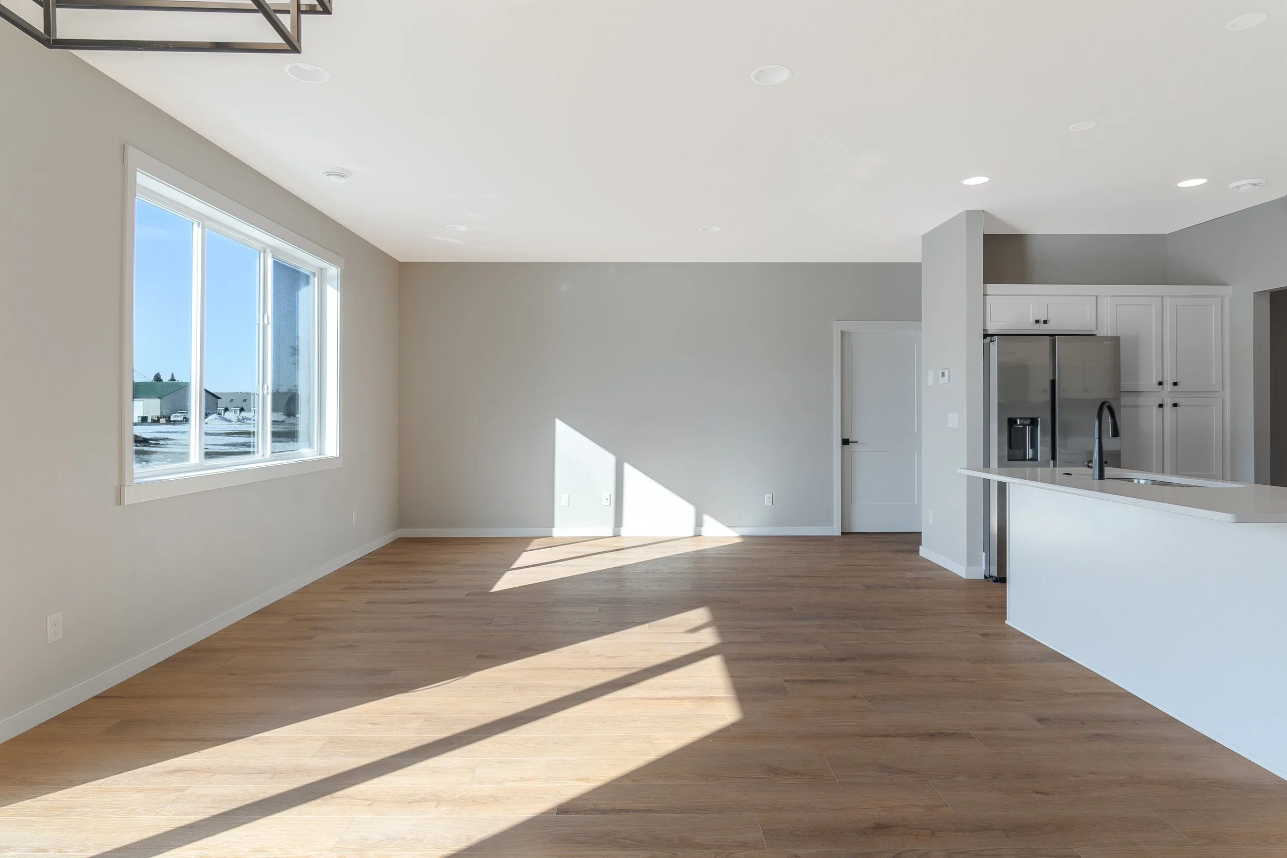 Empty living room with large window, hardwood floors, and open kitchen with white cabinets and stainless steel refrigerator.