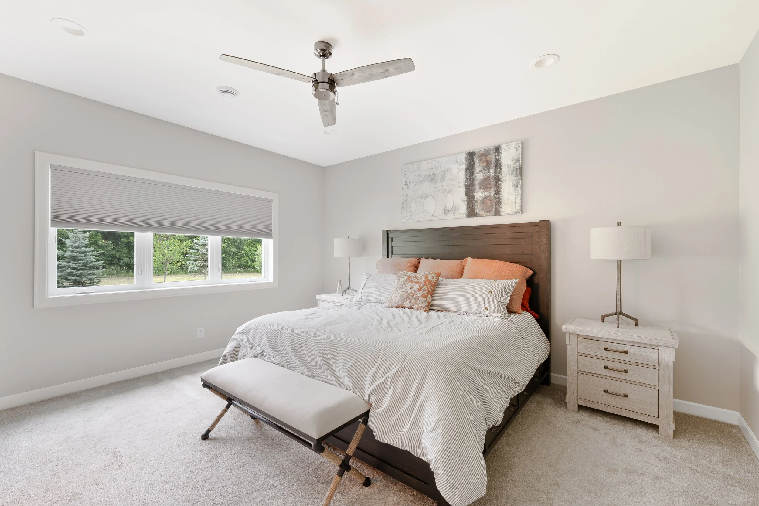 A bedroom with a large bed, pillows, a bedside table with a lamp, and a window with a view of trees outside.