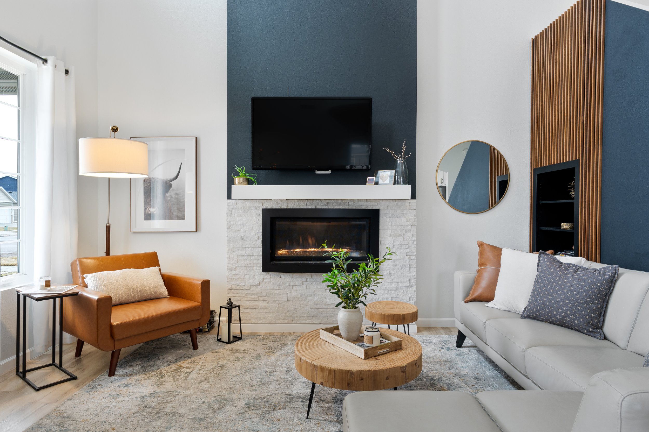 Modern living room with white and dark blue walls, a white brick fireplace, a mounted flat-screen TV, brown leather armchair, white sofa with gray and brown pillows, round wooden coffee table, potted plant, and decorative items.