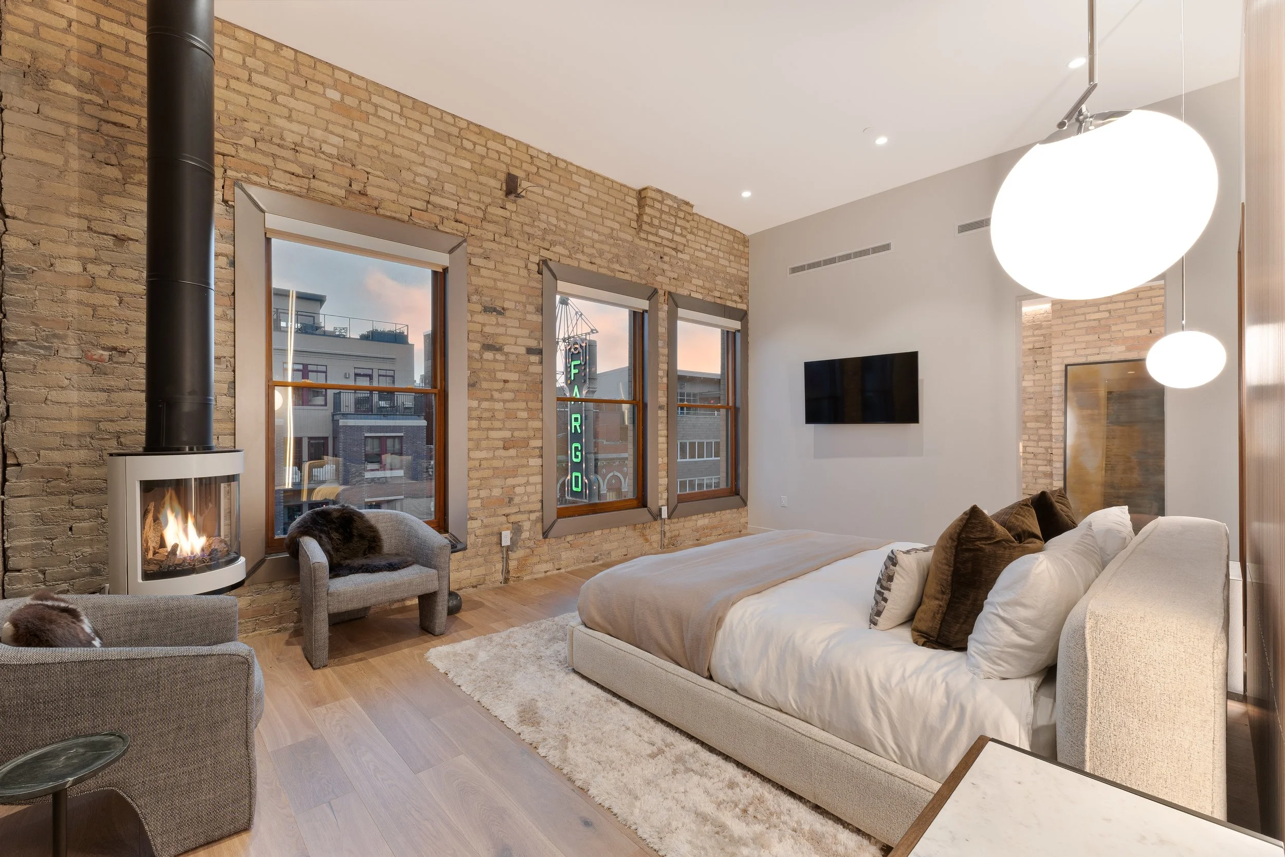 Modern bedroom with exposed brick wall, large window, fireplace, and wall-mounted TV. Bed with white bedding and pillows, cozy chairs with blankets, and contemporary light fixtures.