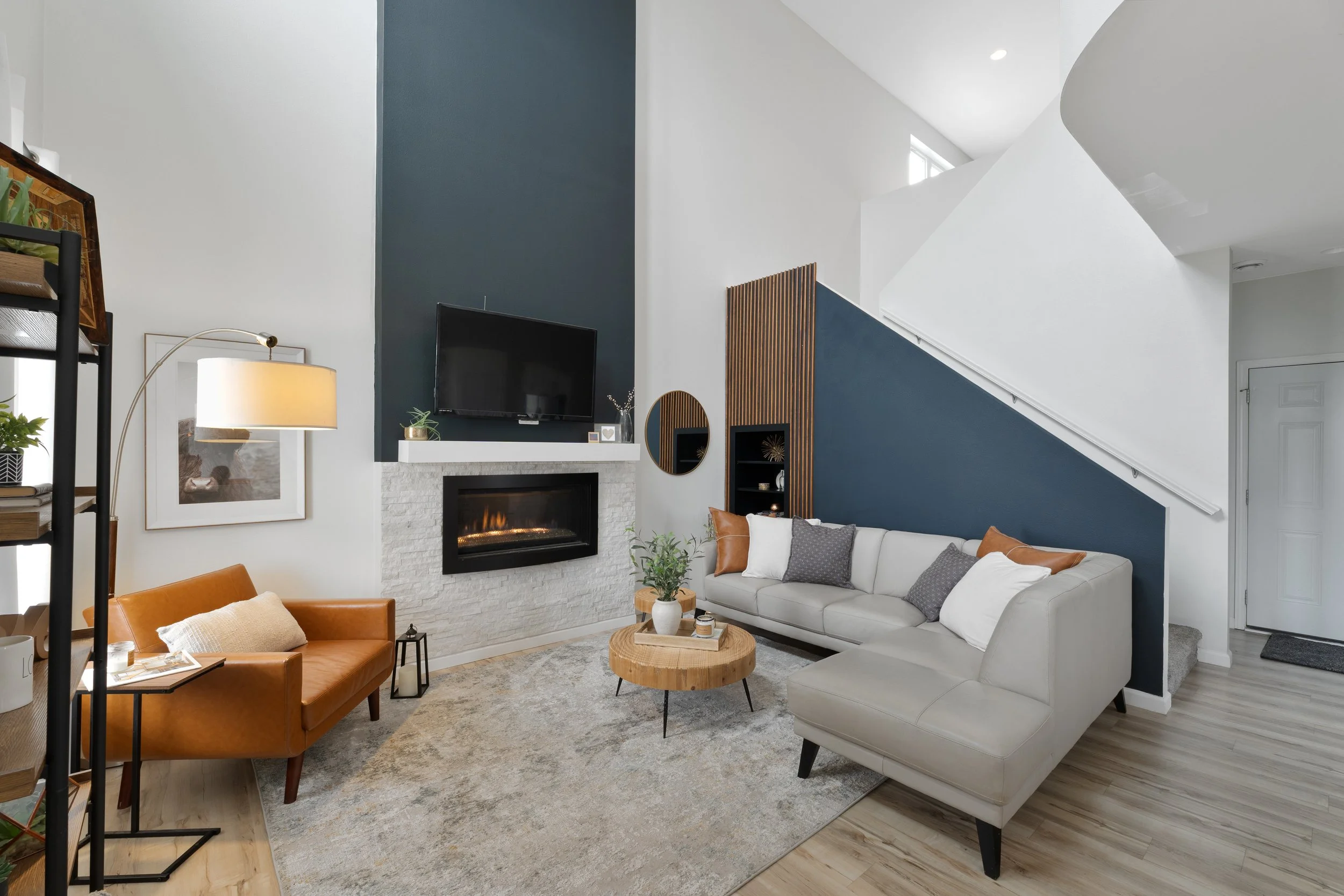 Living room with a white sofa, a brown leather chair, a round wooden coffee table, a fireplace with a TV above, and geometric decor elements, all with modern design.