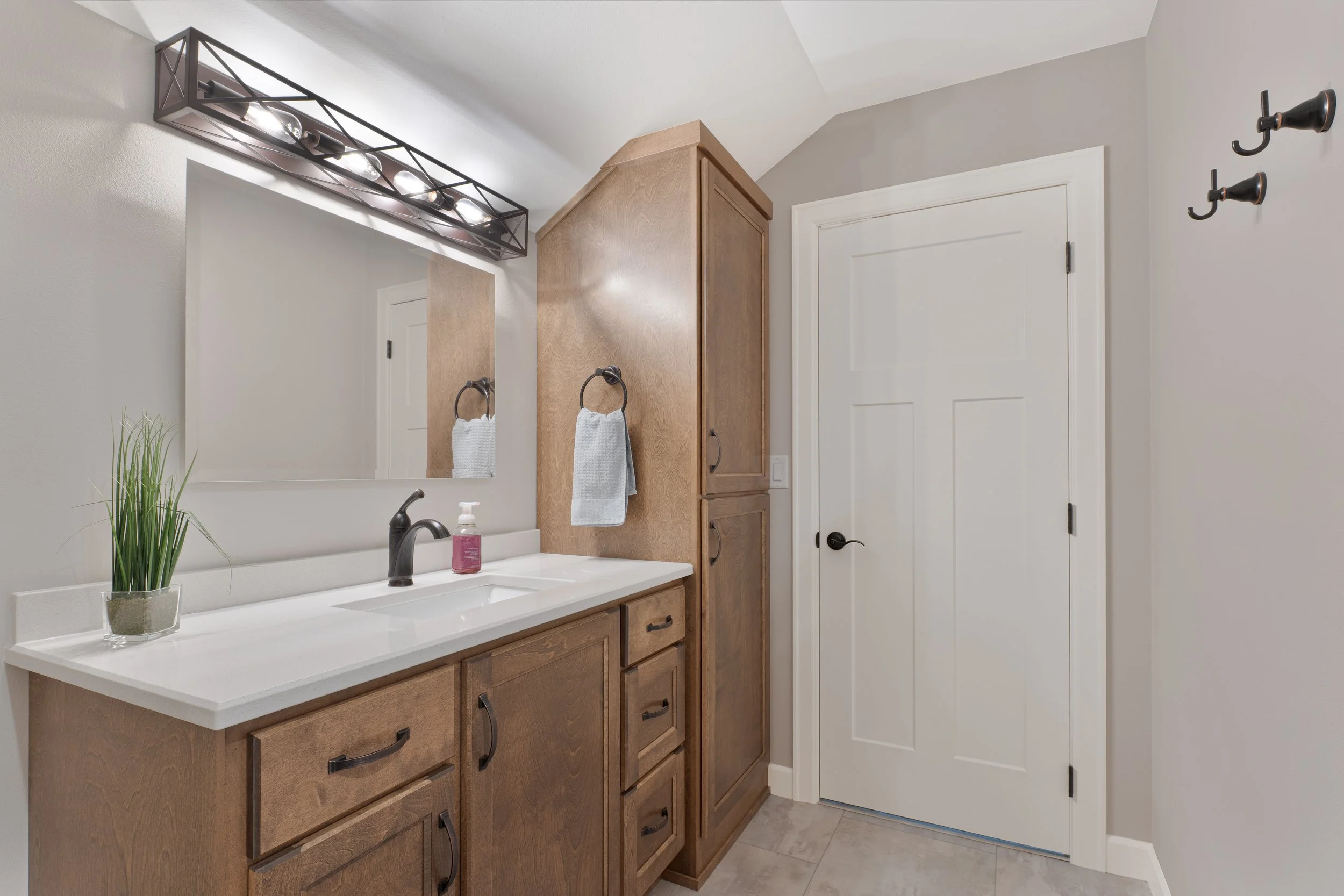 Bathroom with wooden vanity, large mirror, potted plant, soap dispenser, light fixture, white door, gray wall, and black fixtures.