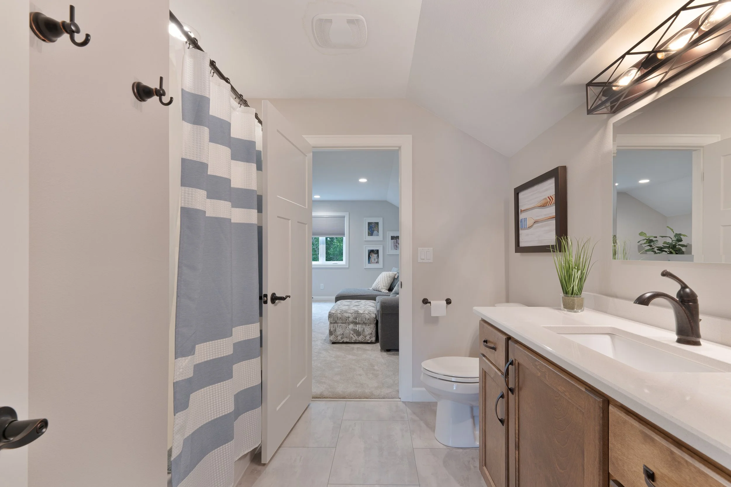 View of a bathroom with a vanity, mirror, and a plant, looking into a bedroom with a gray sofa and framed pictures, near a window.