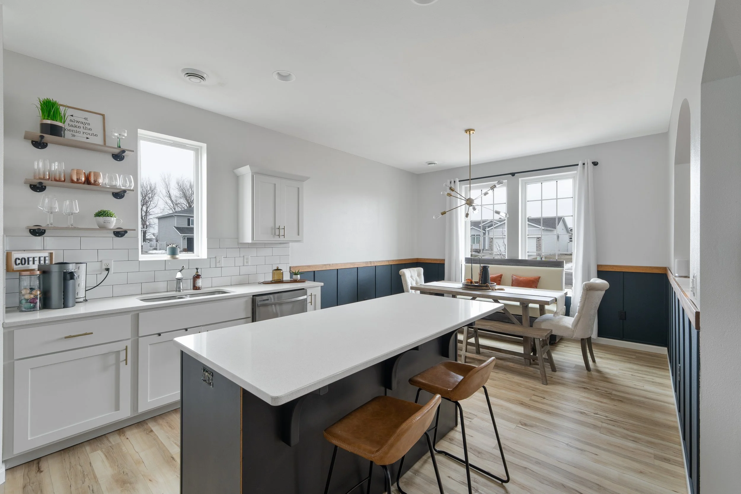 Modern kitchen and dining area with white cabinets, dark blue wainscoting, a white island, and a dining table with chairs and bench, natural light from large windows.