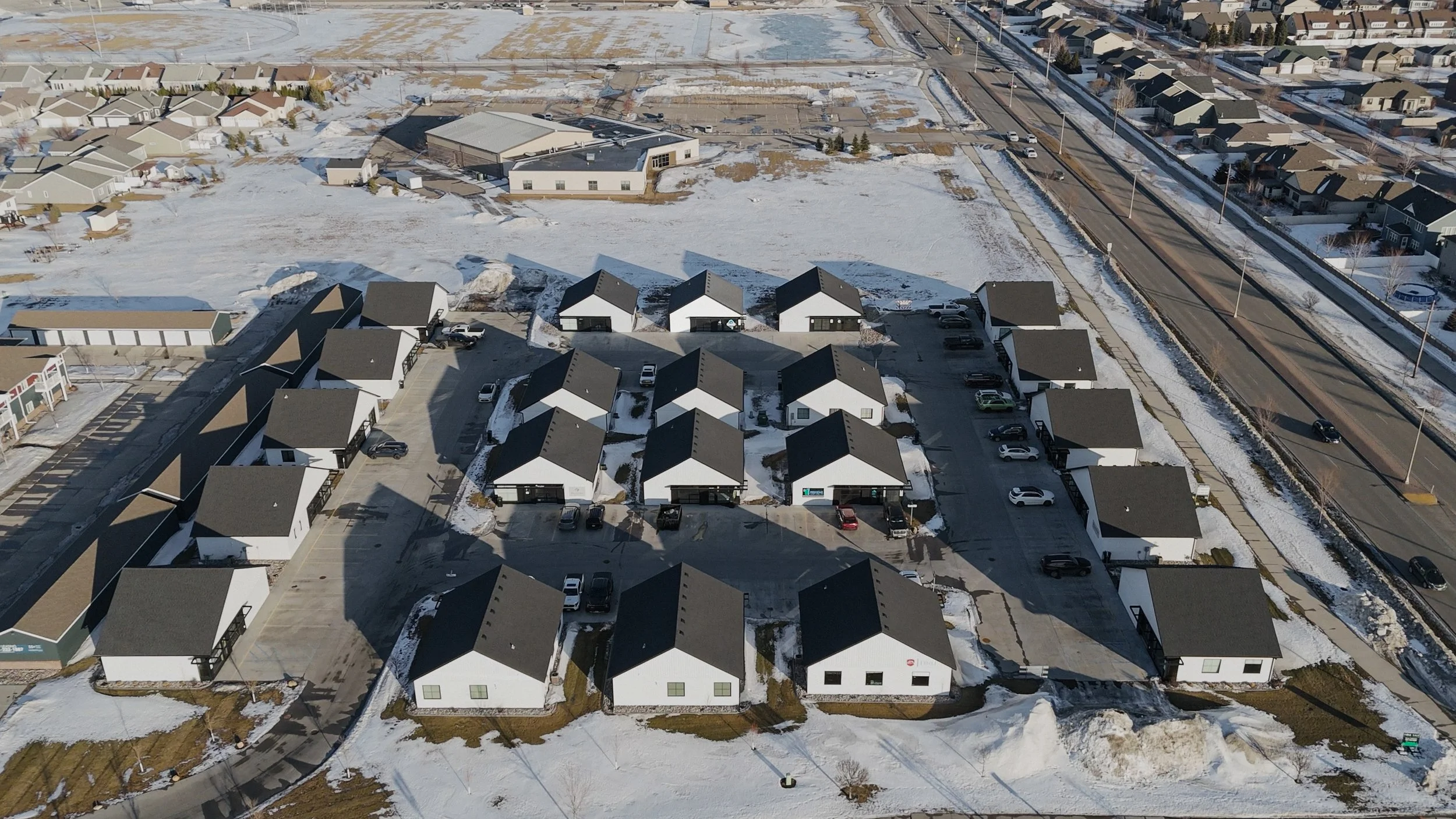 Aerial view of a residential neighborhood with snow-covered ground, houses with dark roofs, parked cars, and adjacent roads in winter.