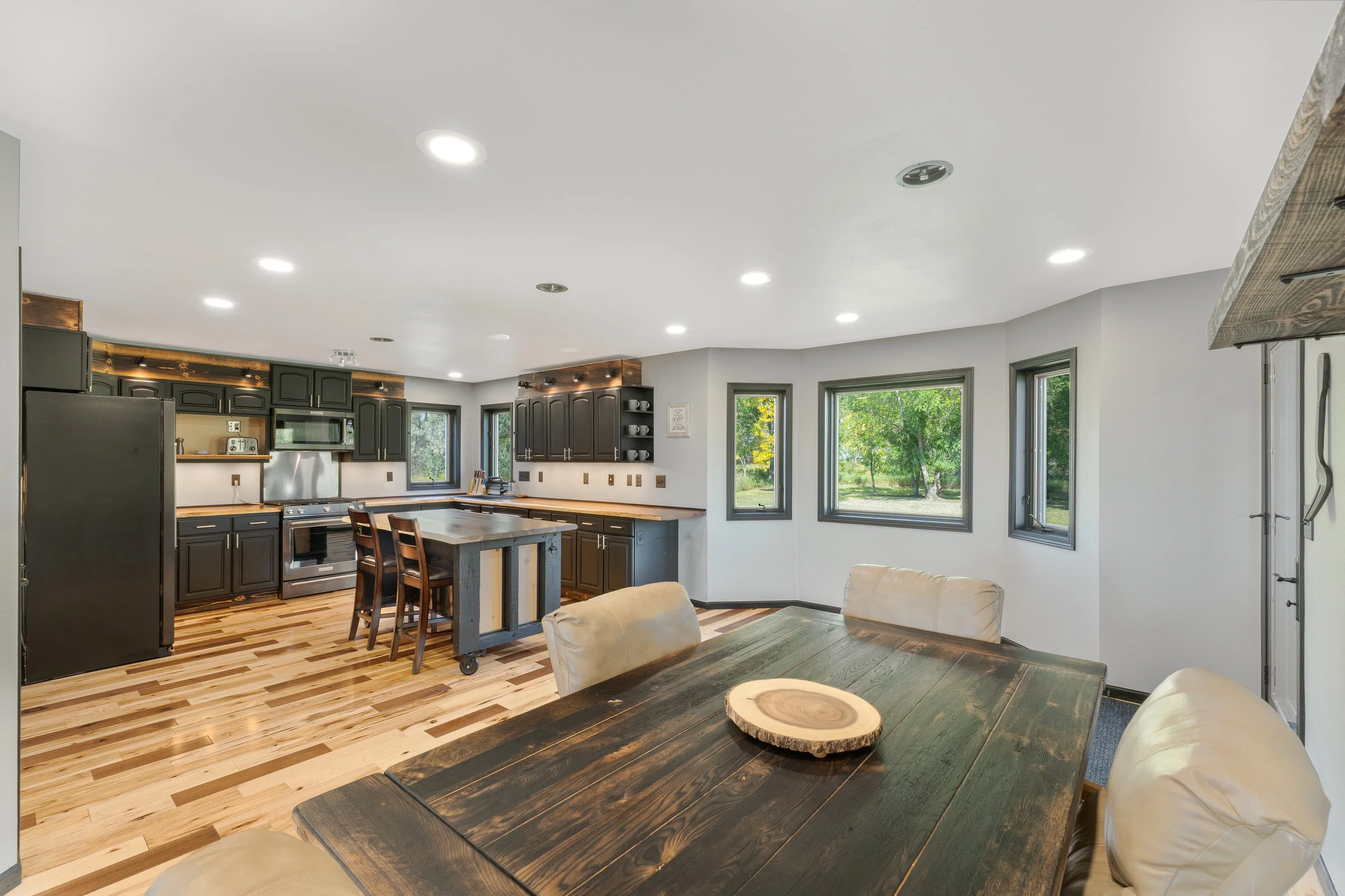 Open concept kitchen and dining area with black cabinets, wooden countertops, a kitchen island with chairs, large windows with greenery outside, and a dark wooden dining table with beige chairs.