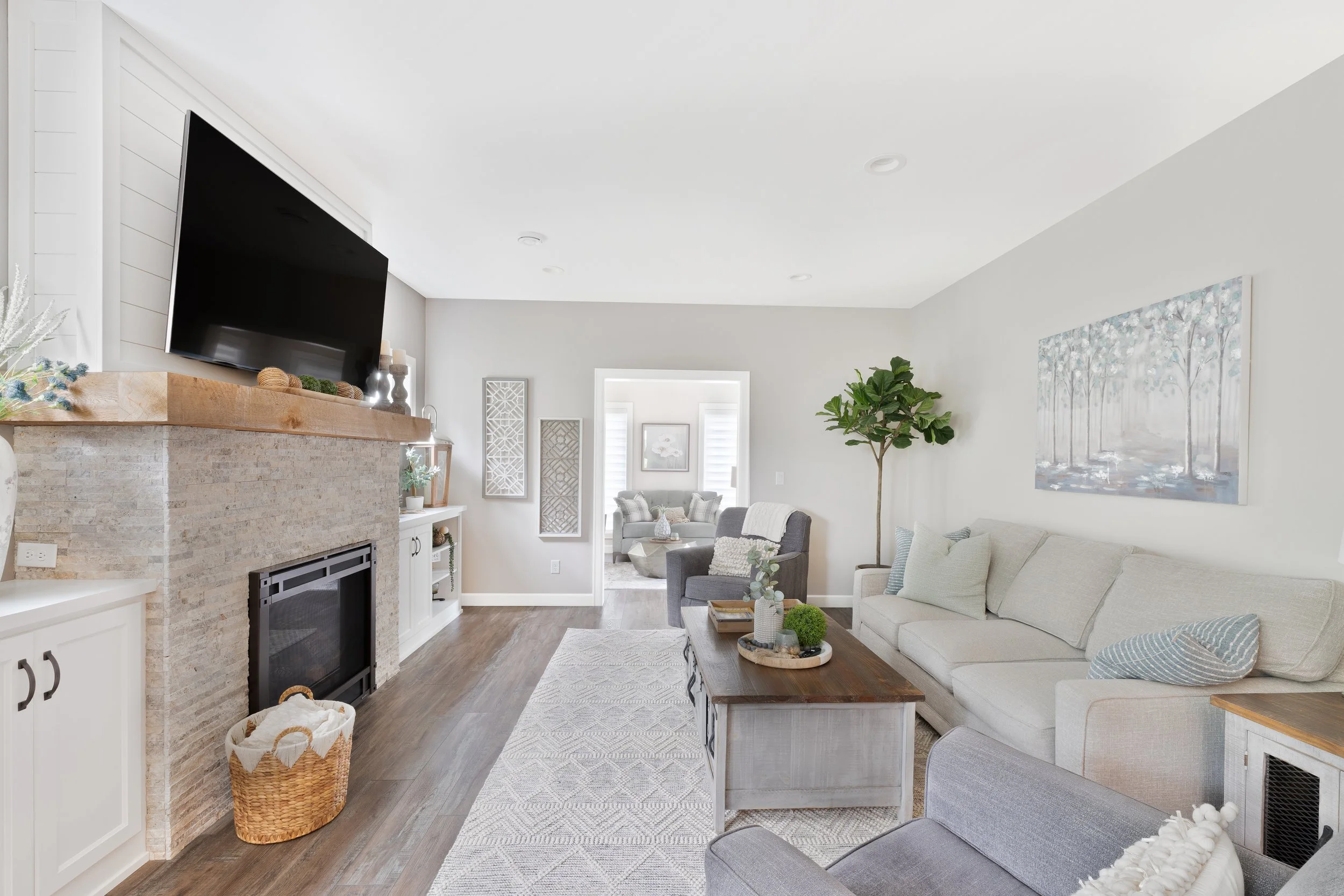 Living room with a beige sectional sofa, gray armchair, wooden coffee table, and a fireplace with a large mounted TV. Decor includes plants, artwork, and pillows, with a view into an adjacent sitting area.