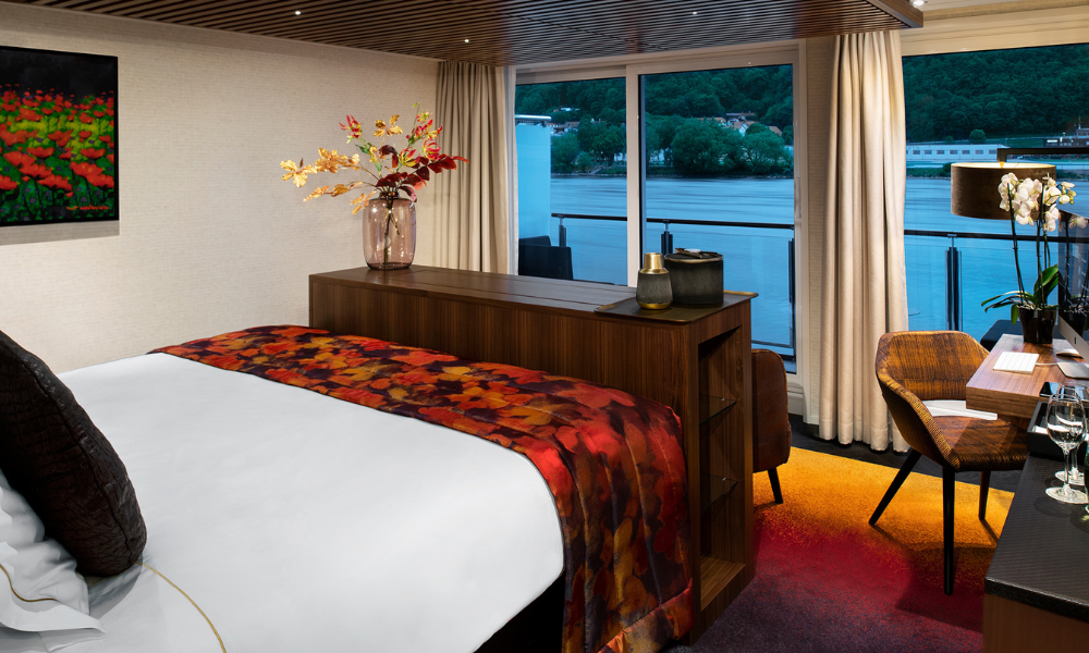 Interior of a modern hotel room overlooking a river with a large bed, floral artwork, a dresser with decorative items, a desk with glasses, and a balcony with a view of the water and trees.