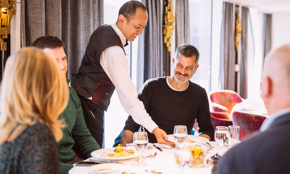 A waiter serving food to a group of people at a restaurant table, with smiles and glasses of water.