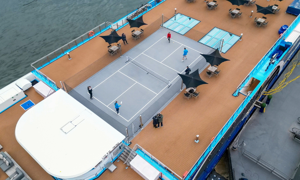 Aerial view of a rooftop deck on a cruise ship with a small tennis court, seating areas with black umbrellas, and people playing tennis.