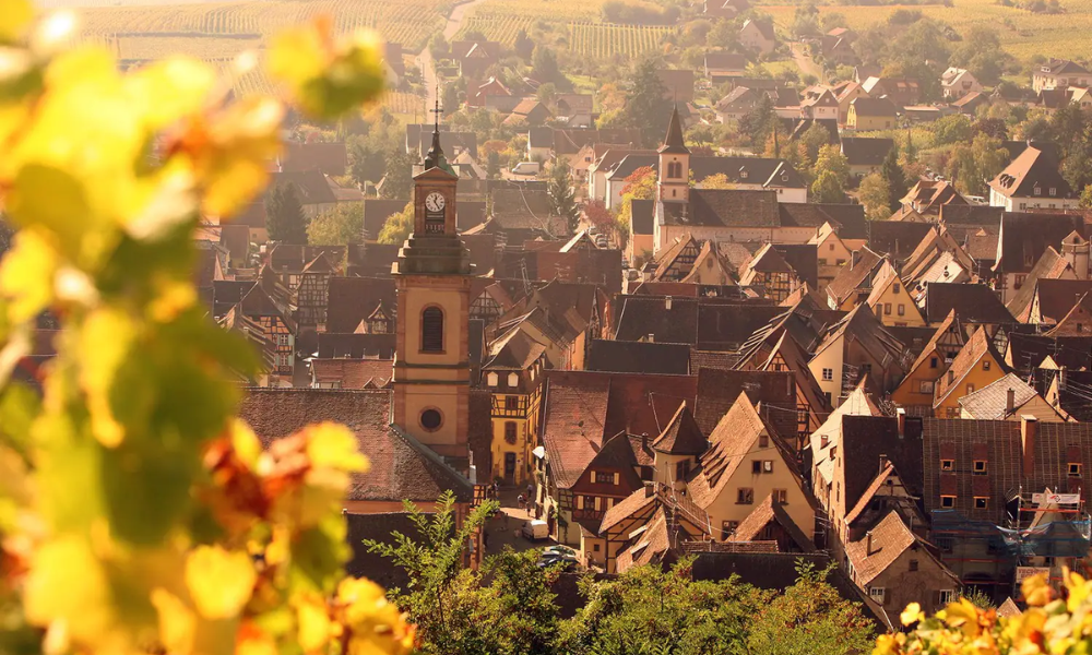 View of a quaint European town with historic churches, surrounded by rolling hills and foliage, during sunset.