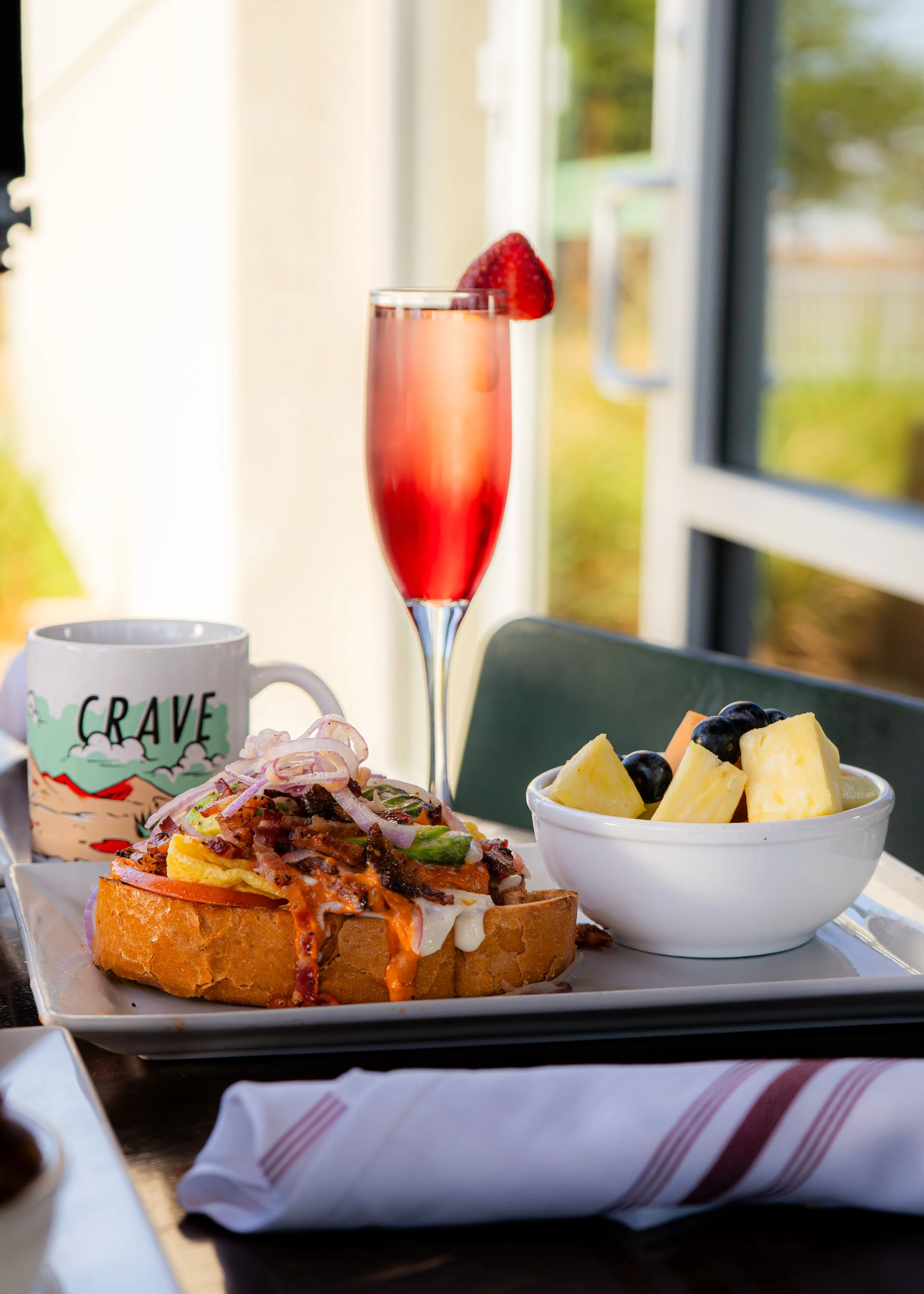 Breakfast serving with a bowl of fruit, a pastry topped with onions and vegetables, a mug, and a glass of strawberry champagne, set on a table near a window.