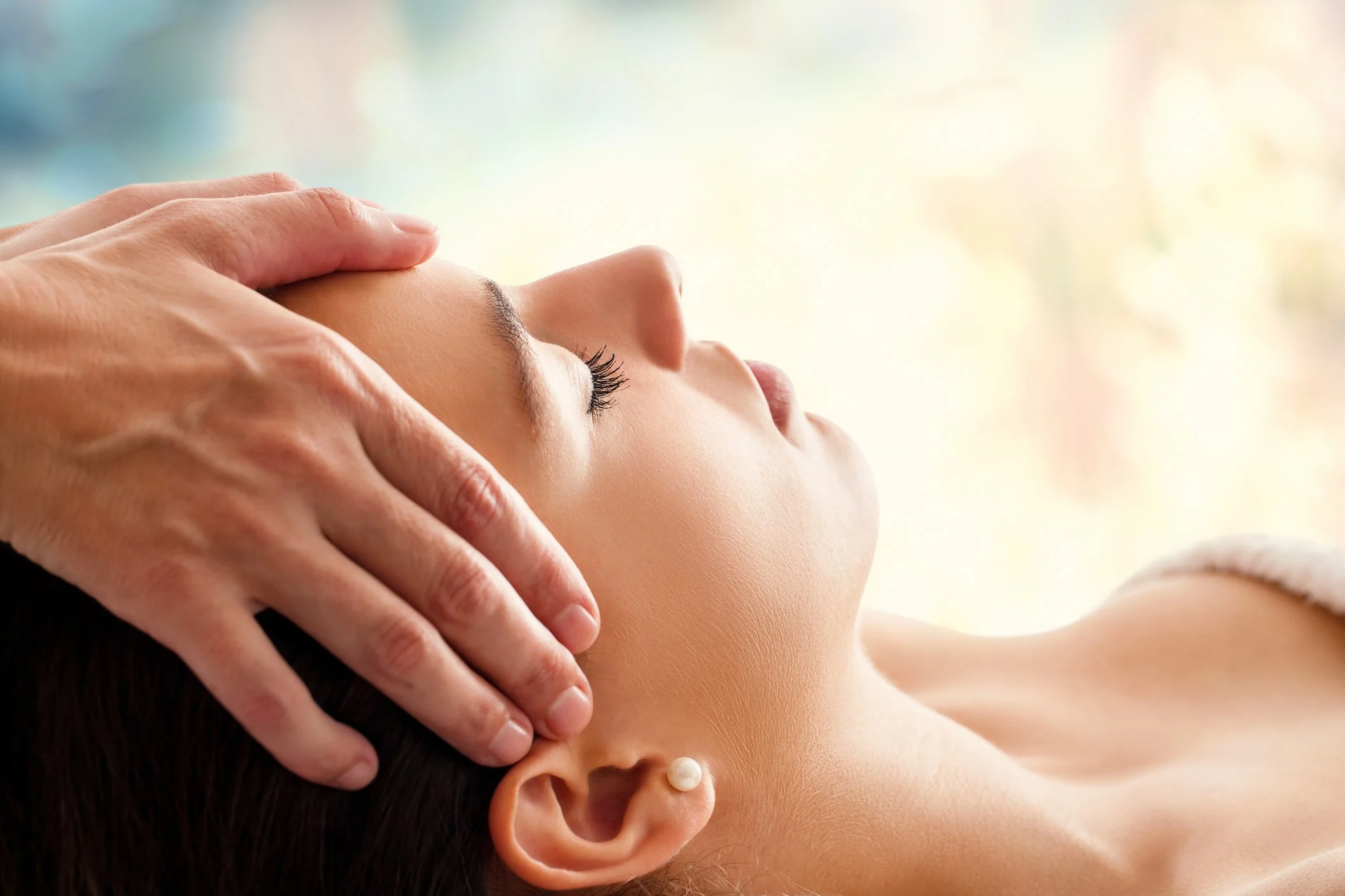 Person receiving a massage or facial treatment, lying down with eyes closed, a hand gently placed on their forehead.