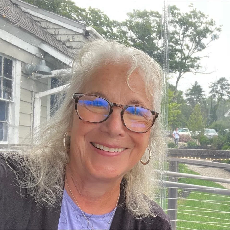 Smiling woman with glasses and earrings sitting outdoors near a window with a building and trees visible in the background.
