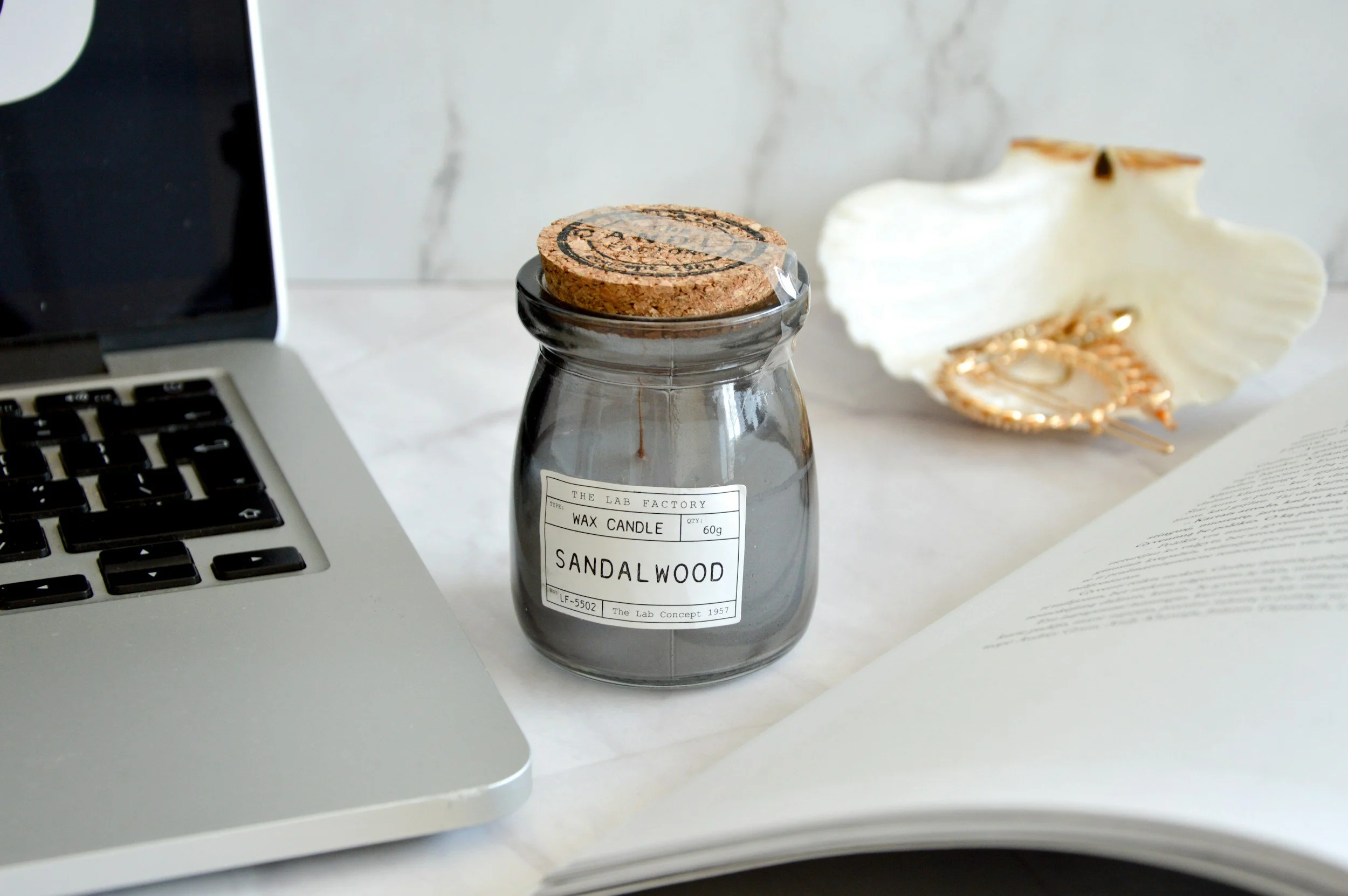 A workspace with a MacBook laptop, a candle labeled "Sandalwood" in a glass jar with a cork lid, a shell-shaped dish with gold jewelry, and an open book on a white surface.