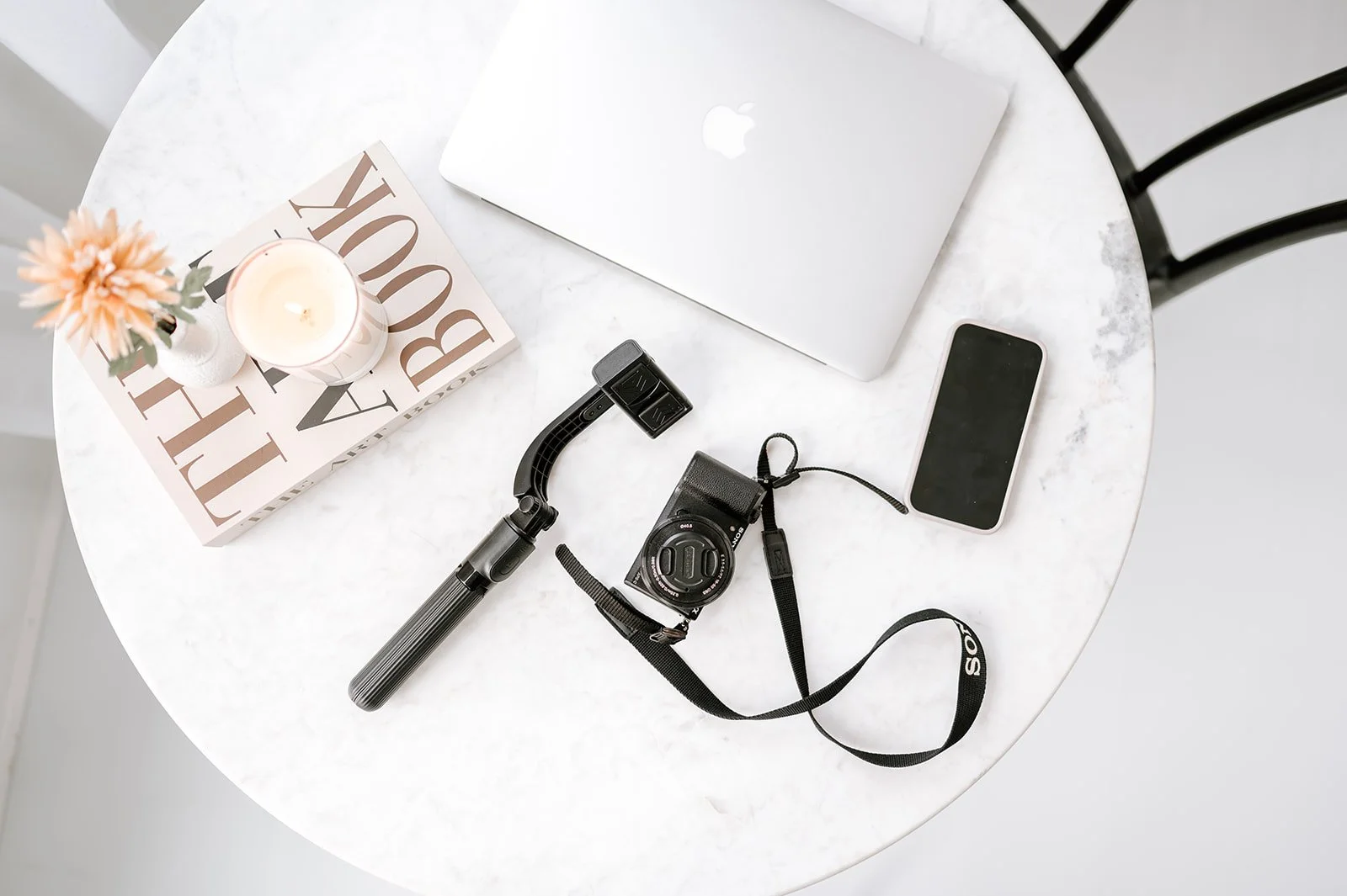 A white round table with a closed Apple laptop, a smartphone, a camera with a strap, a selfie stick, a magazine with candles and a flower on top of it.