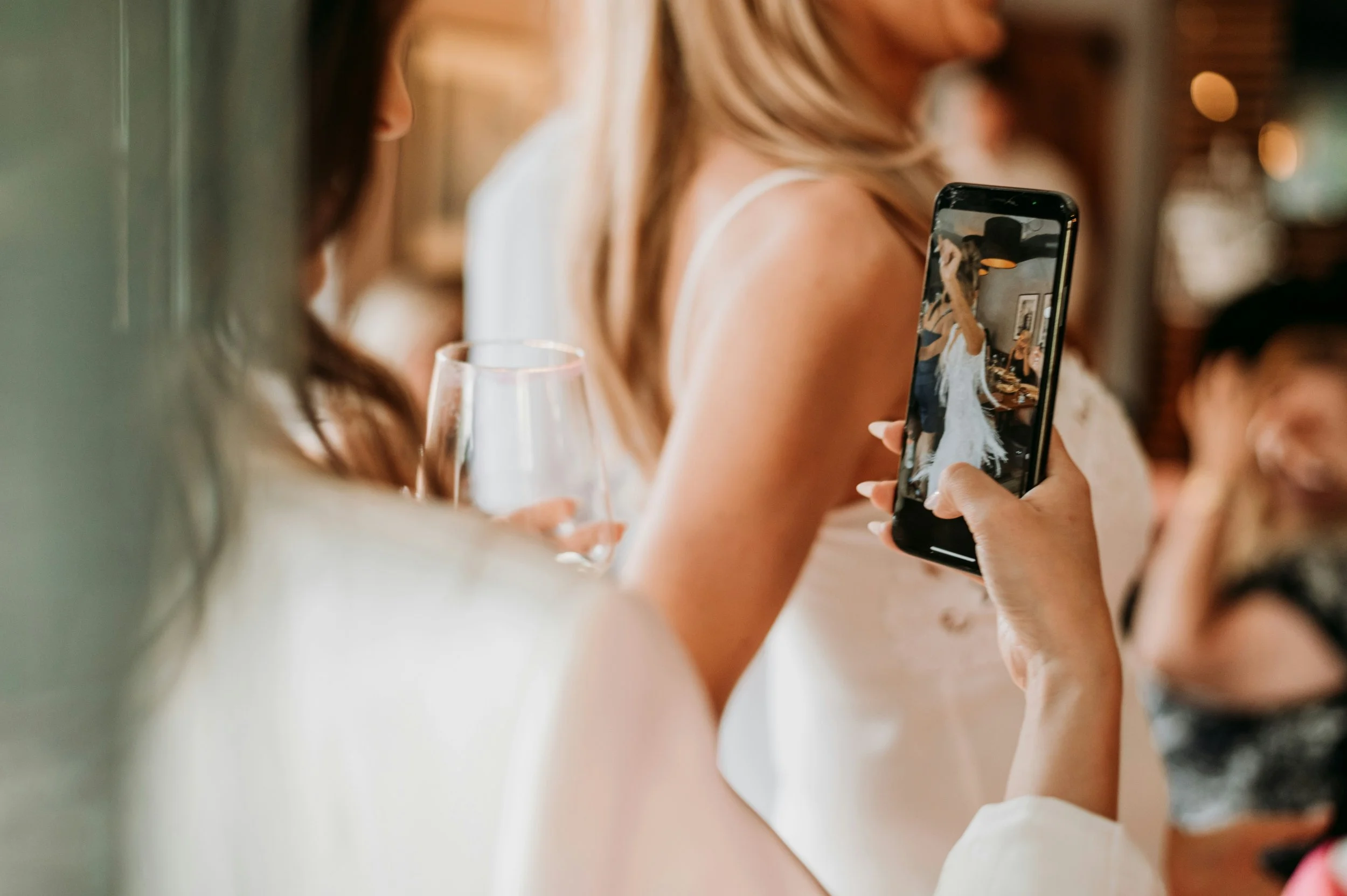 A woman is taking a photo of another woman in a white dress at a social event, with a wine glass in her hand.