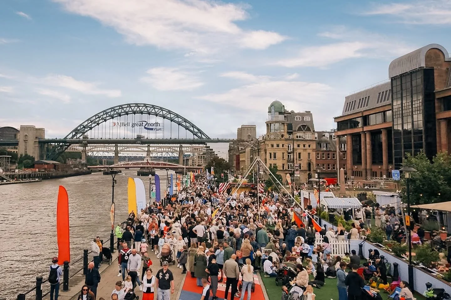 Crowd of people gathered along a riverfront walkway with colorful flags and tents, city buildings and a large bridge in the background.