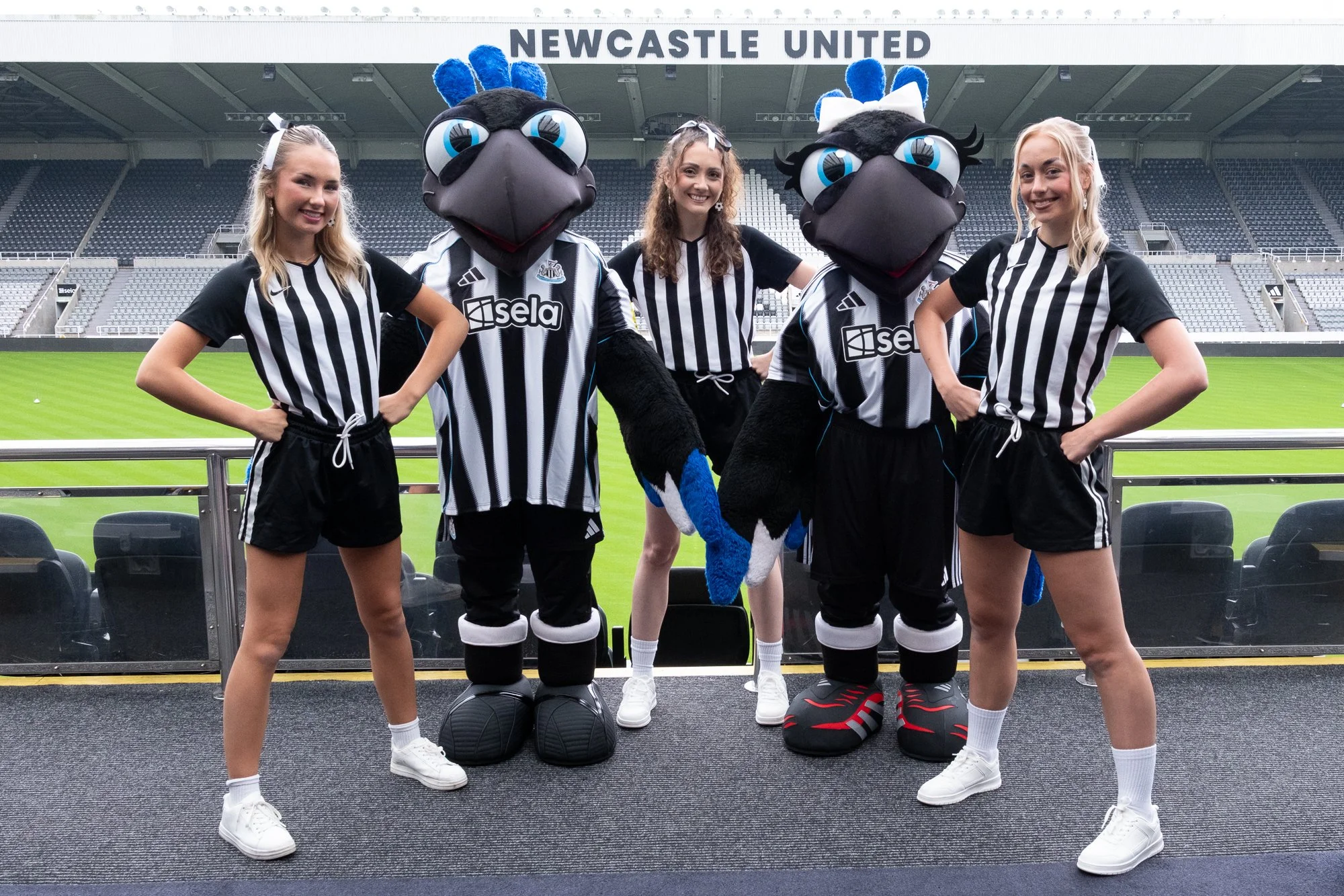 Group of young women and two zookeepers dressed as Newcastle United mascots standing on a balcony at a football stadium, smiling for a photo.