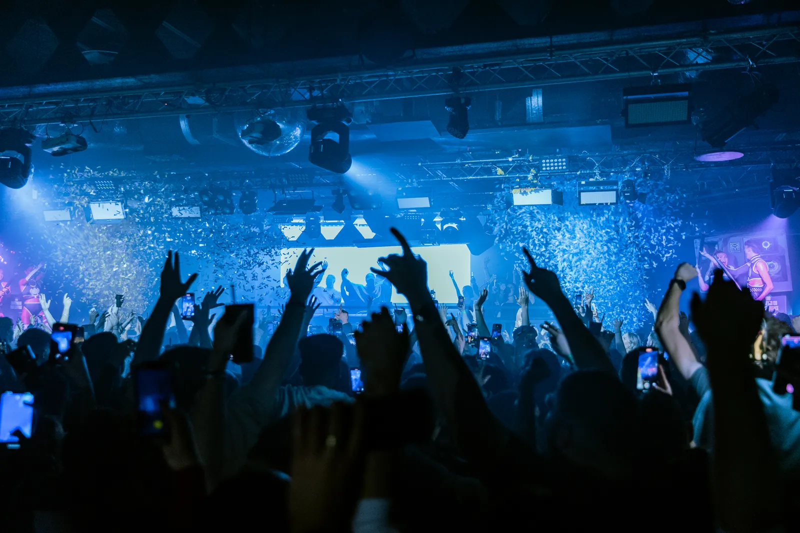 Crowd at a concert or nightclub showing raised hands, phone screens, and a stage with performers amid confetti and colorful lighting.