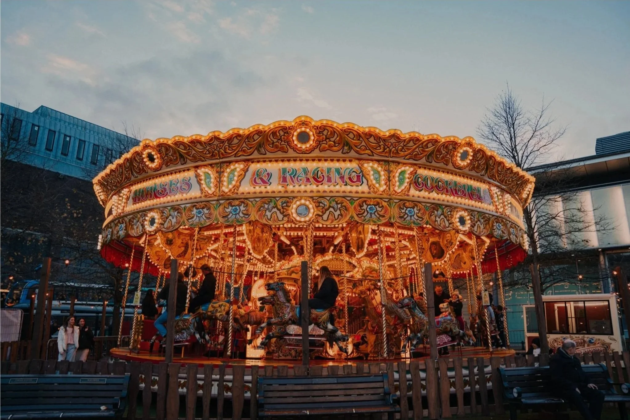 A brightly lit vintage carousel with ornate decorations, including floral patterns and gold accents, is situated outdoors in an urban area during early evening. People are riding the carousel and walking nearby, with modern buildings and leafless trees in the background.