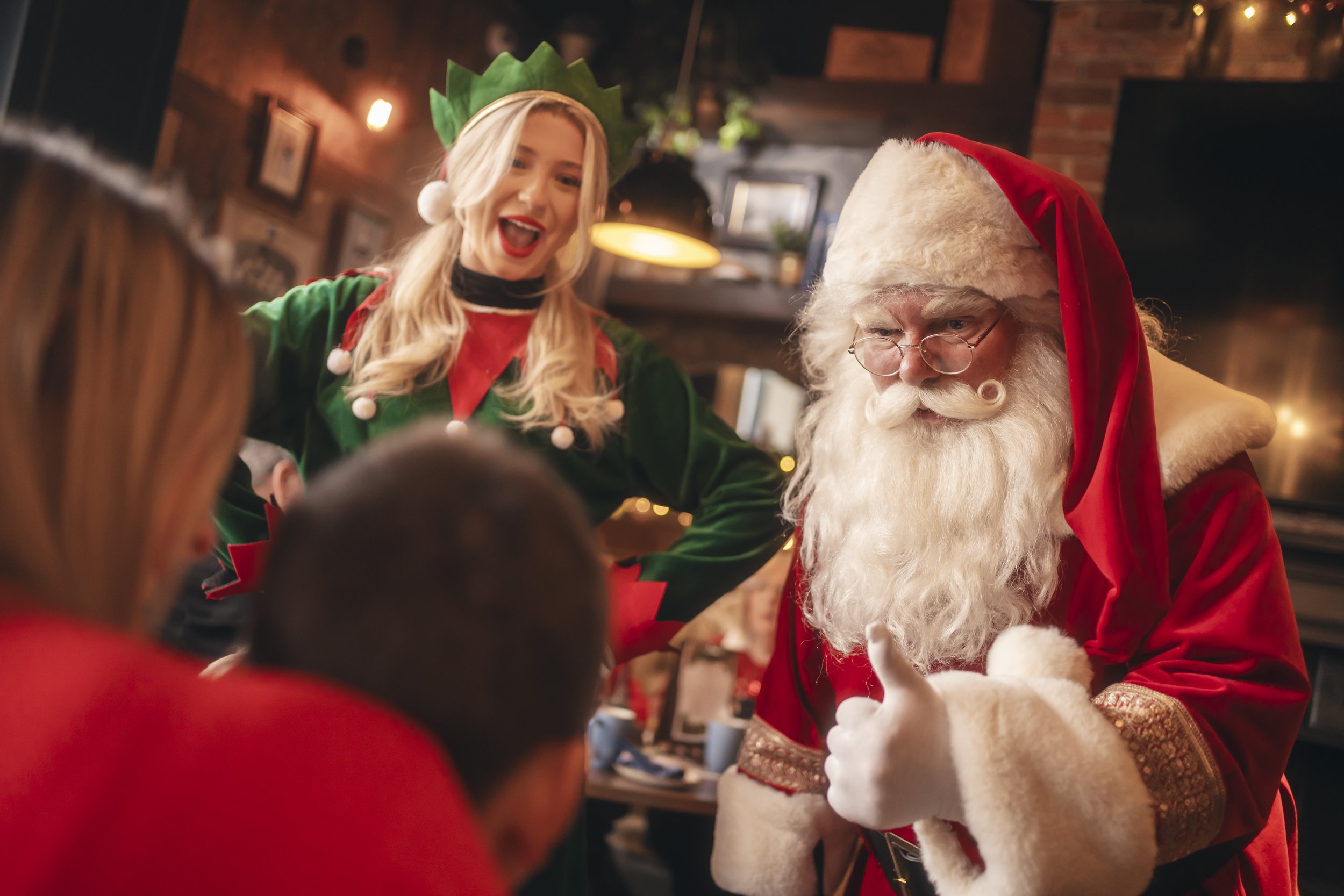 A woman dressed as an elf with a green hat and red lipstick is smiling while interacting with Santa Claus, who is wearing a red hat with white fur trim, glasses, and a red and white costume, at a festive holiday gathering.
