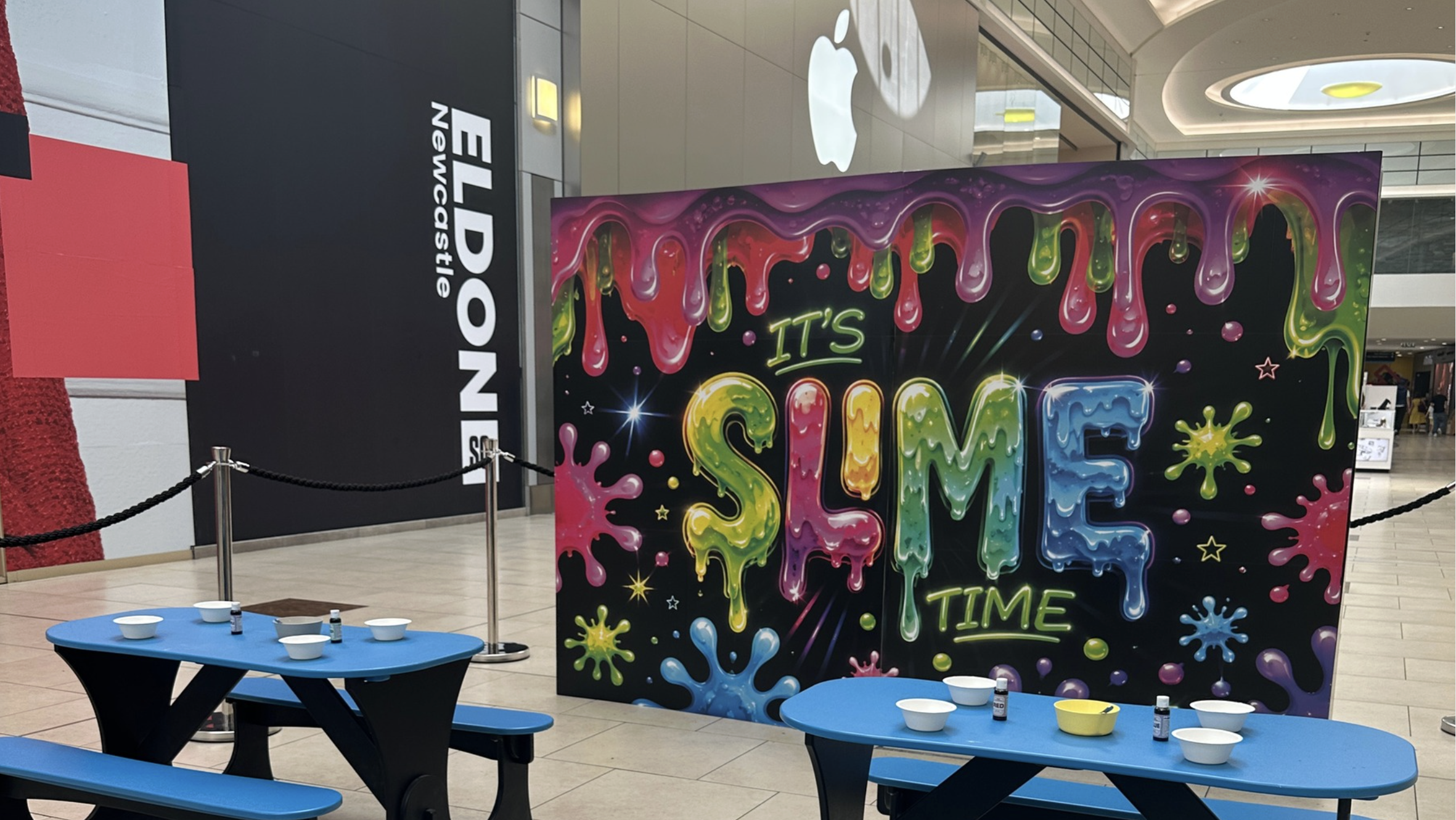 Colorful sign with the words "IT'S SUMMER TIME" in neon and paint splatter design, displayed in a shopping mall.