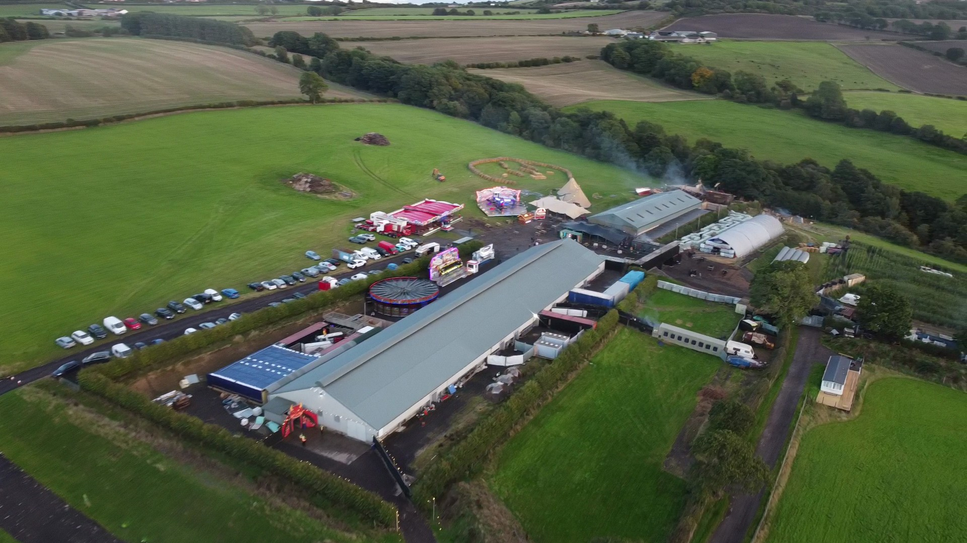 Aerial view of a fairground with amusement rides, tents, and parking lot surrounded by green fields and farmland.