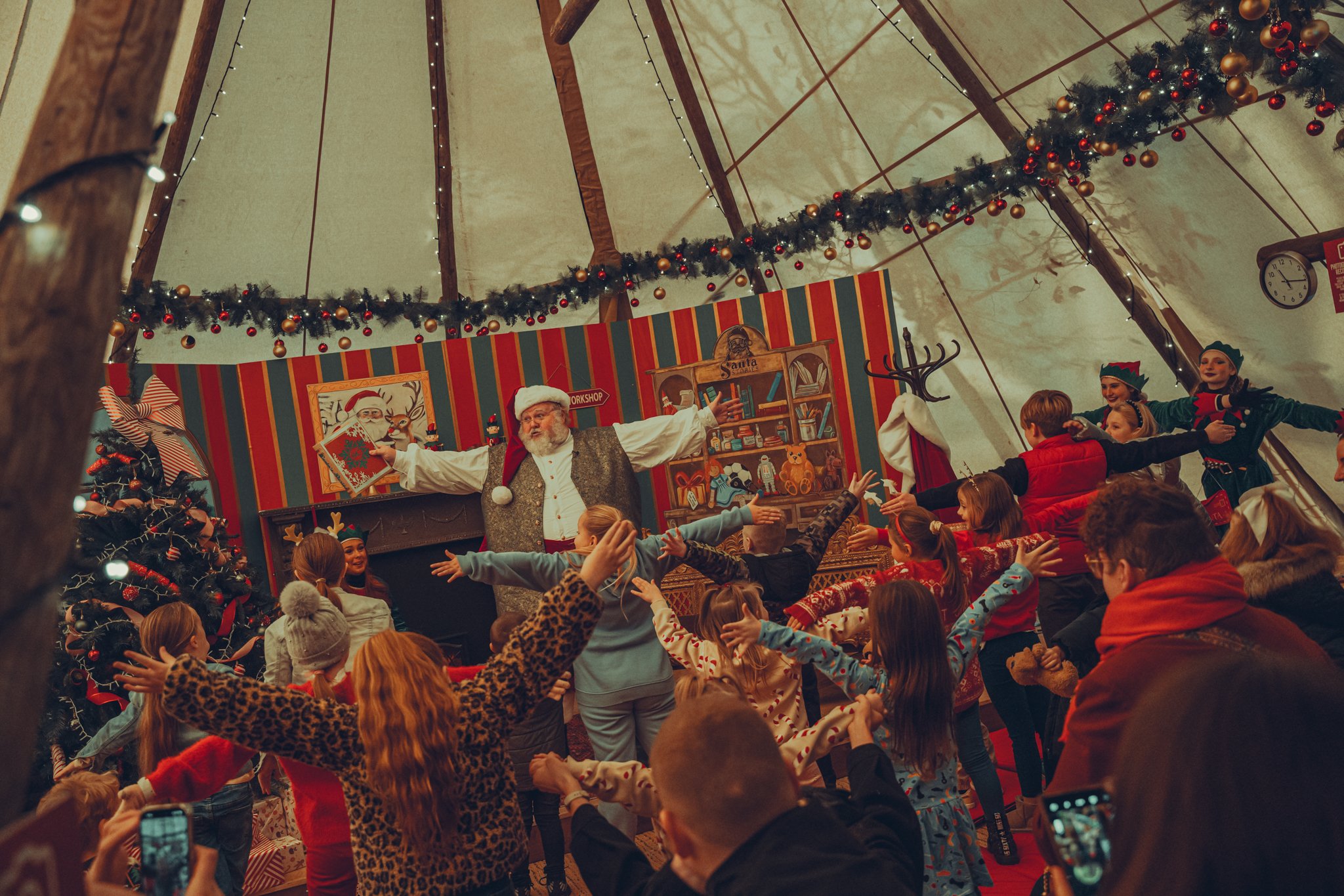Children and Santa Claus holiday performance inside a decorated tent with Christmas garland and a Christmas tree.