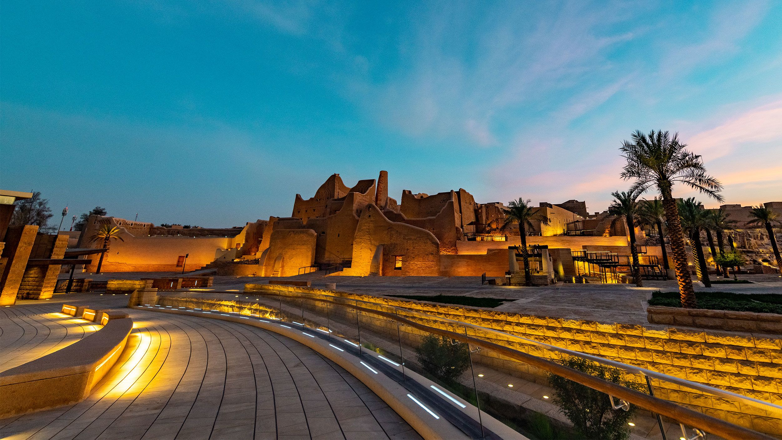A historic adobe fortress with palm trees in a modern landscaped area during dusk, illuminated with warm lighting.