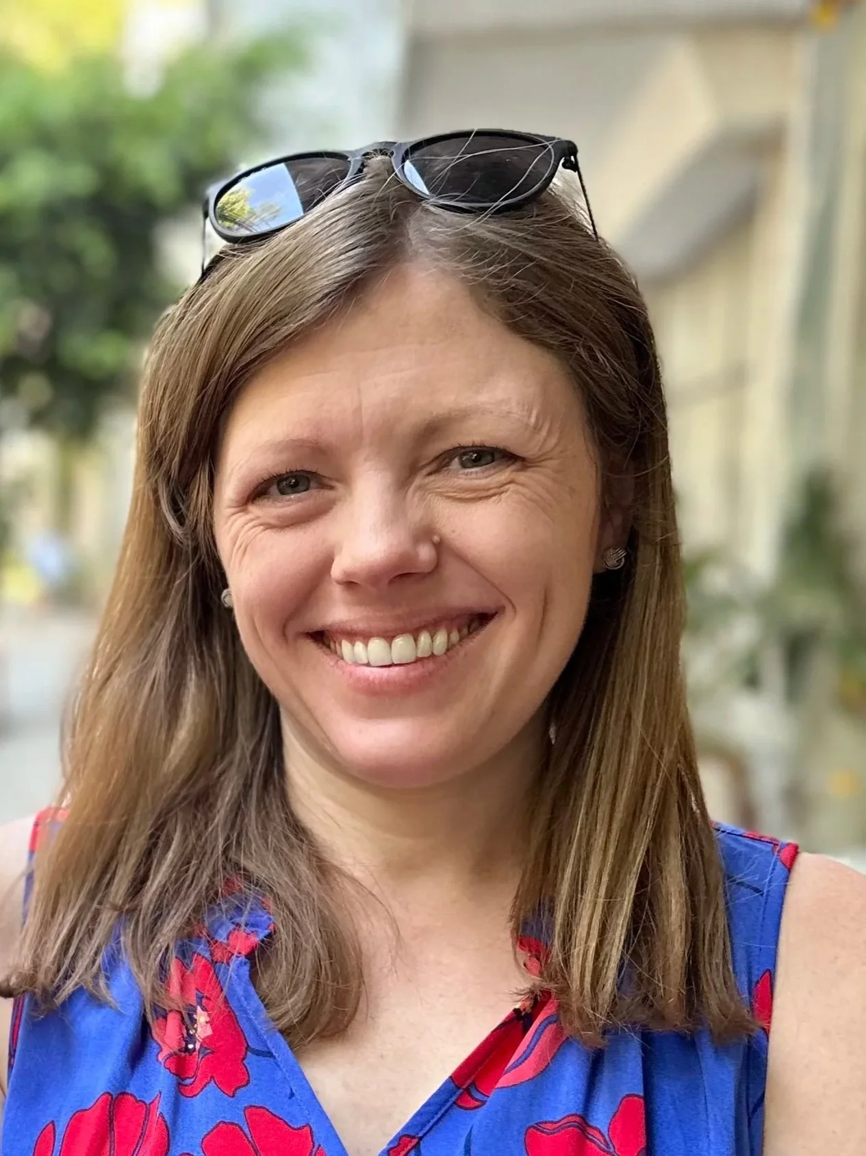 A smiling person with short hair, wearing a septum piercing, earrings, a gray shirt, and orange shorts, standing outdoors against a background of green foliage.