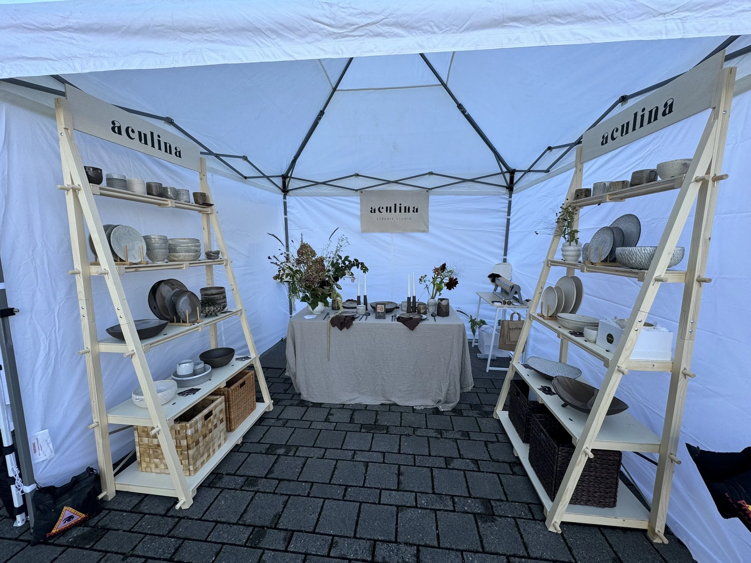 Booth displaying ceramic dishes and bowls at an outdoor market, with shelves on the left and right, a table with vases and flowers in the middle, under a white tent.