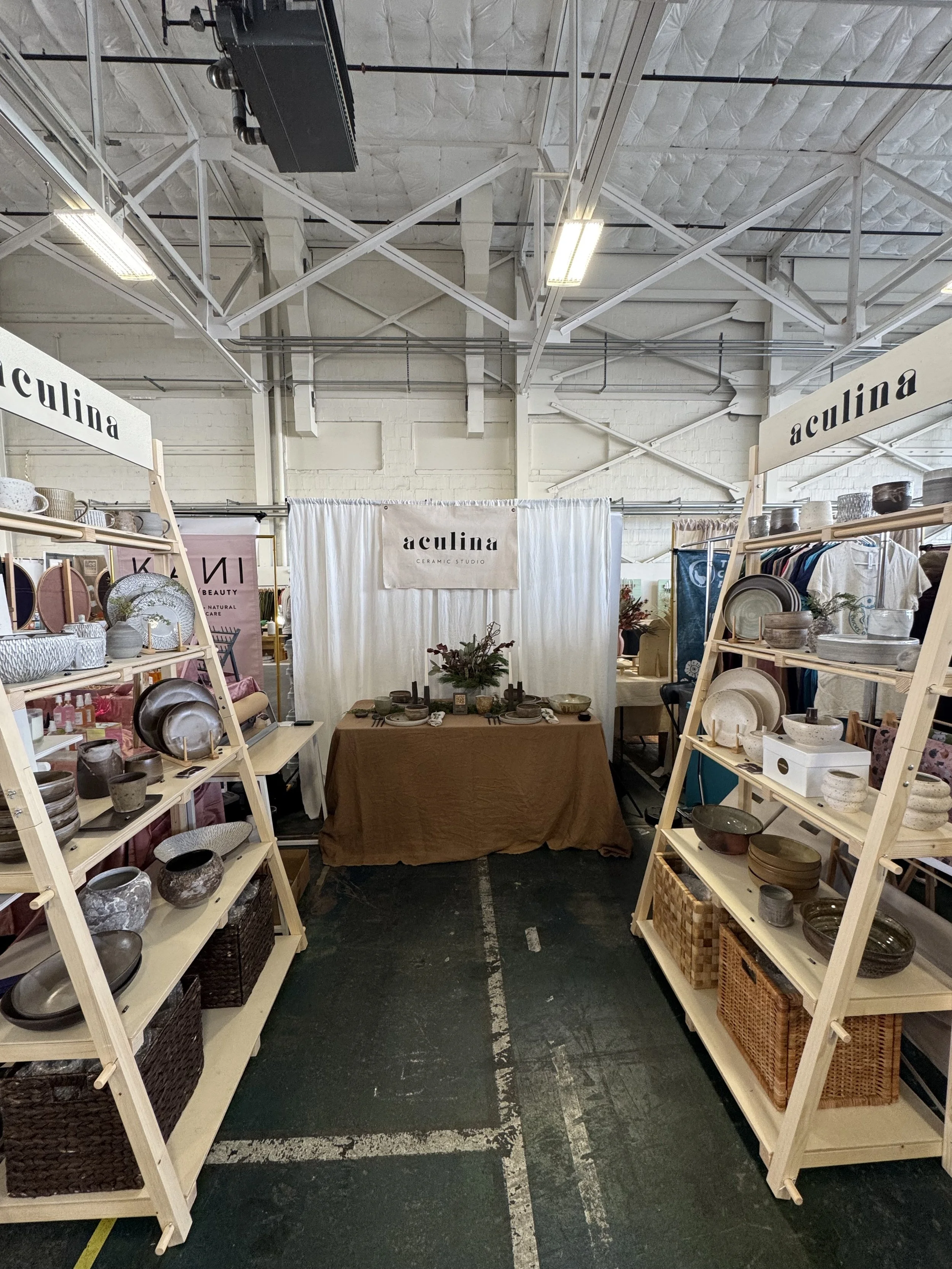 Ceramic pottery display booth at a craft market with shelves holding bowls, plates, and cups, and a table in the center decorated with plants and ceramics, under industrial ceiling lighting.