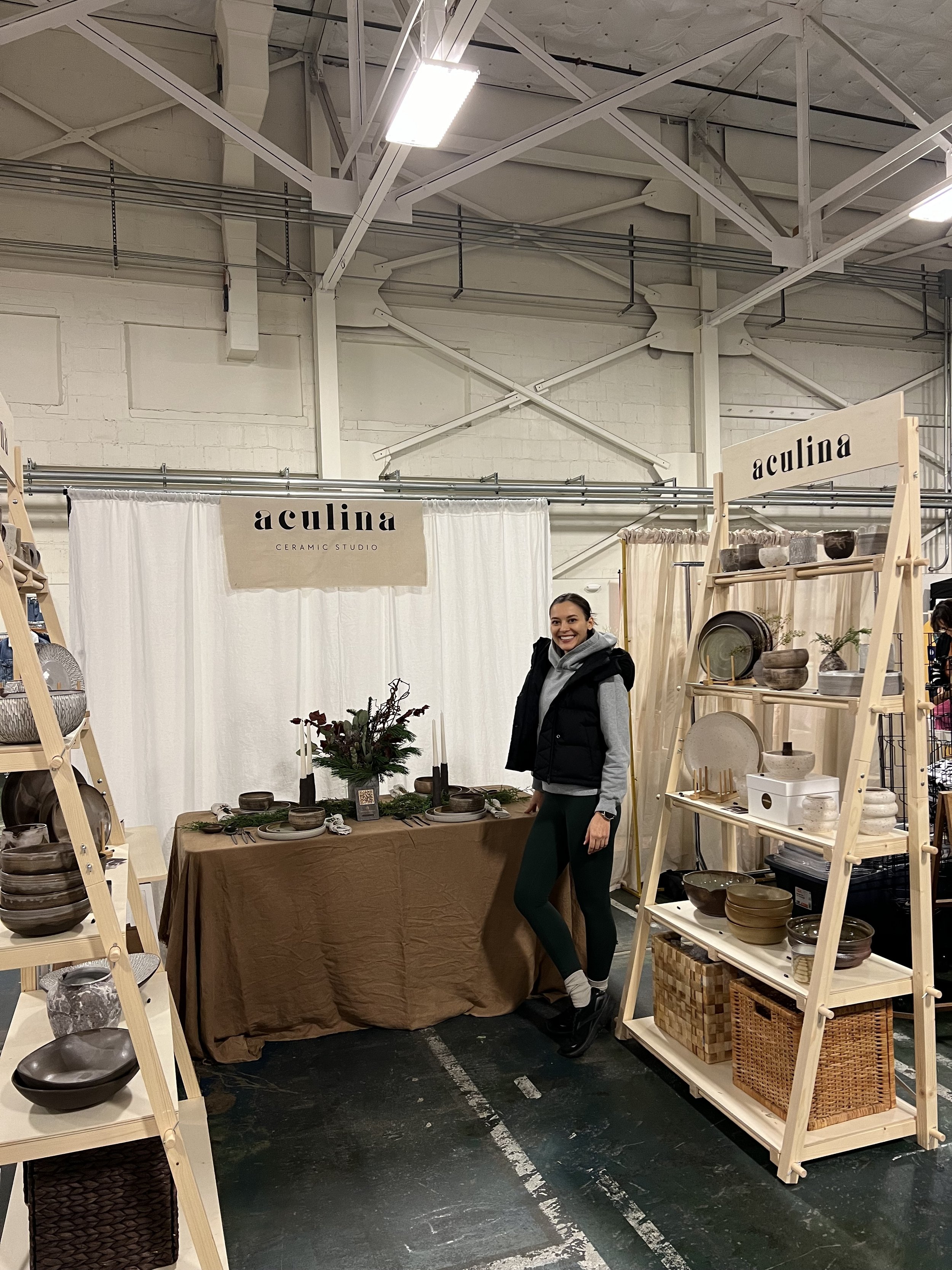 A woman standing at a ceramic booth display with shelves of pottery and a table with dinnerware, at an indoor market or craft fair.