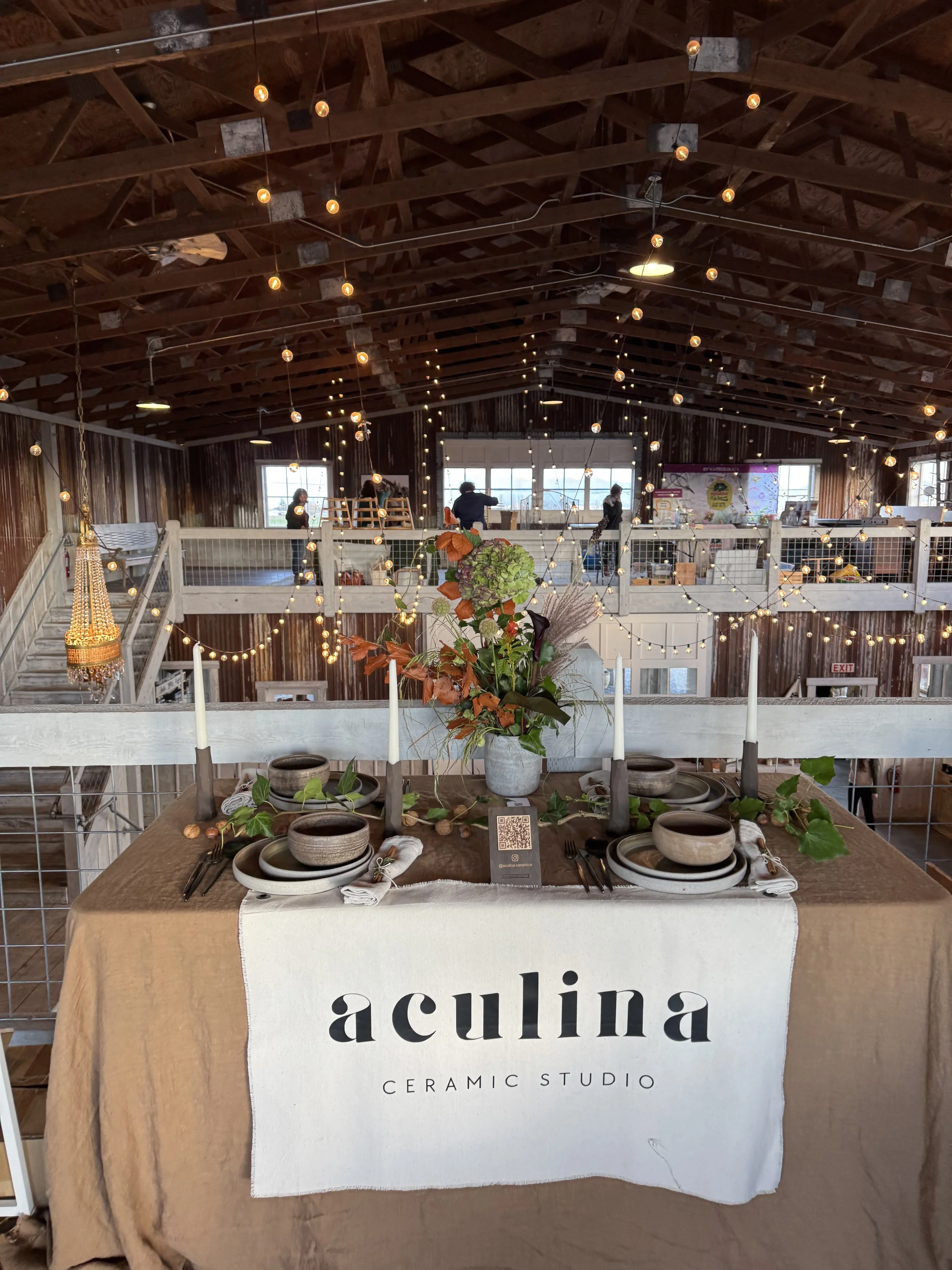 Table set for dining at Aculina Ceramic Studio with a beige tablecloth, candles, florals, and tableware. The background features warm string lights and a rustic wooden interior with multiple levels, large windows, and people browsing.