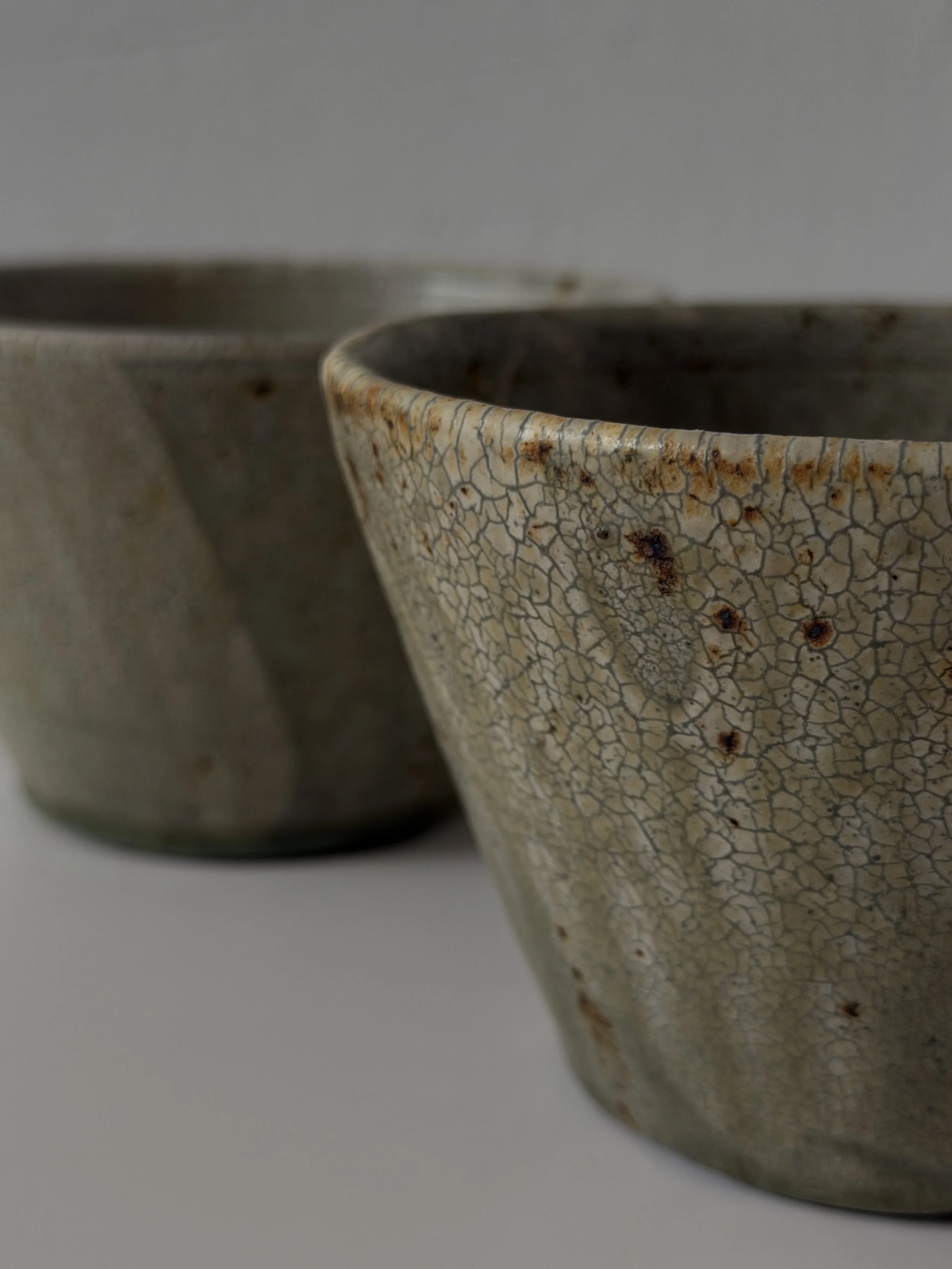 Close-up of two rustic ceramic bowls with crackle glaze and earthy tones, placed on a plain surface.