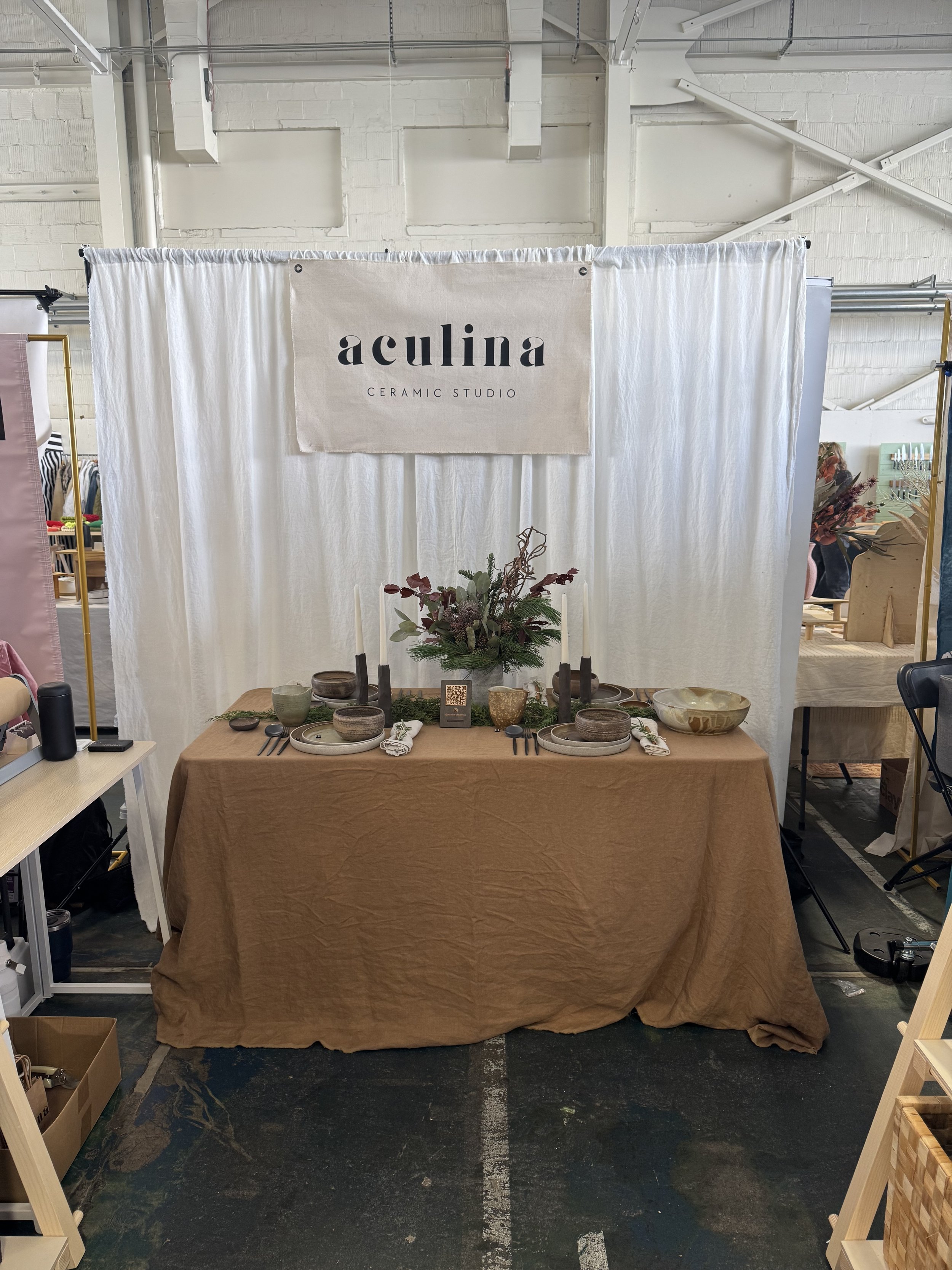 Display booth with a white backdrop and a sign reading 'aculina CERAMIC STUDIO.' In front, a table covered with a brown cloth holds a floral arrangement, candles, bowls, plates, cutlery, and napkins, set up for a ceramic showcase.