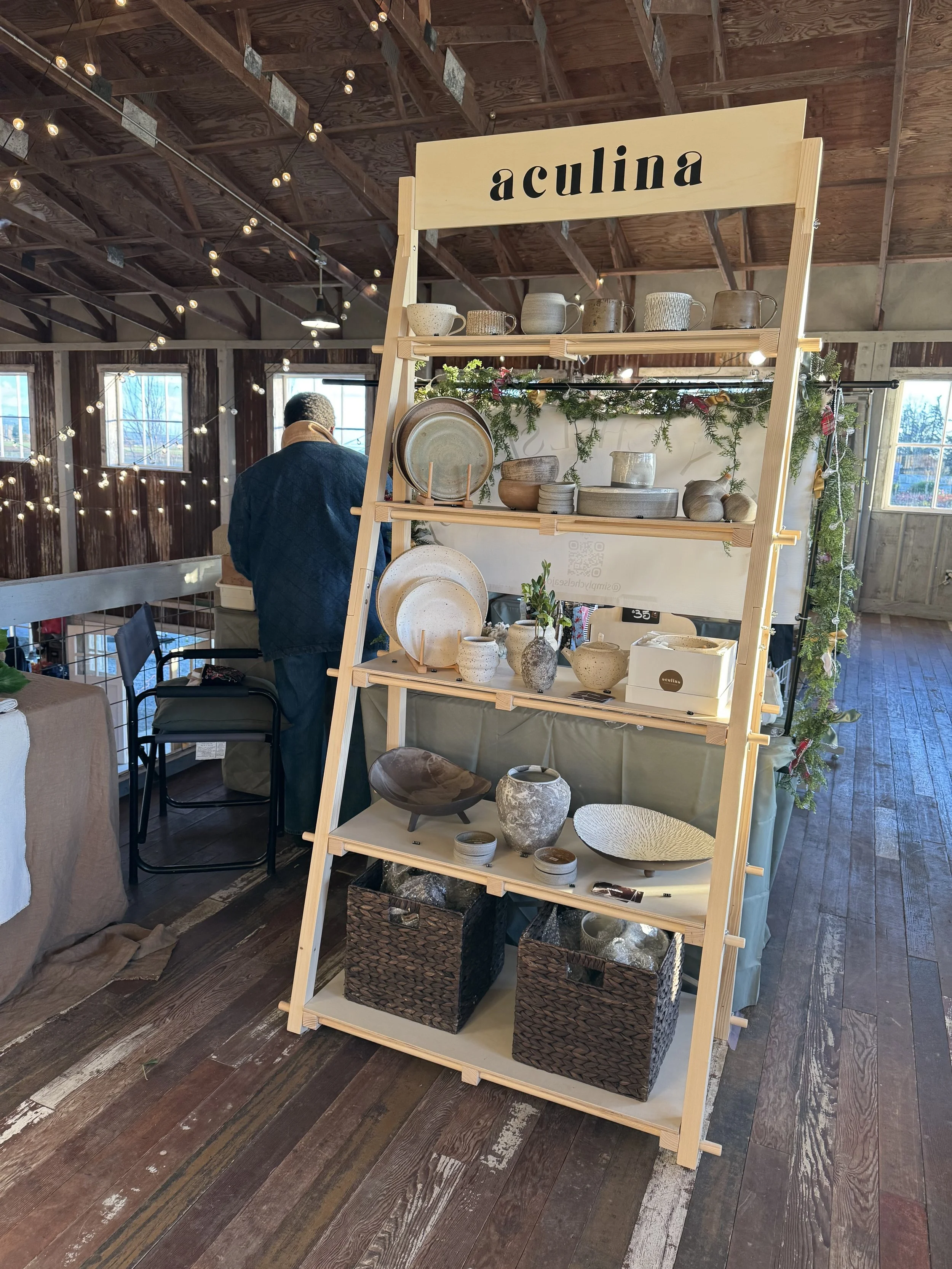 Display stand with handmade pottery and ceramics at a craft fair inside a rustic barn with string lights.