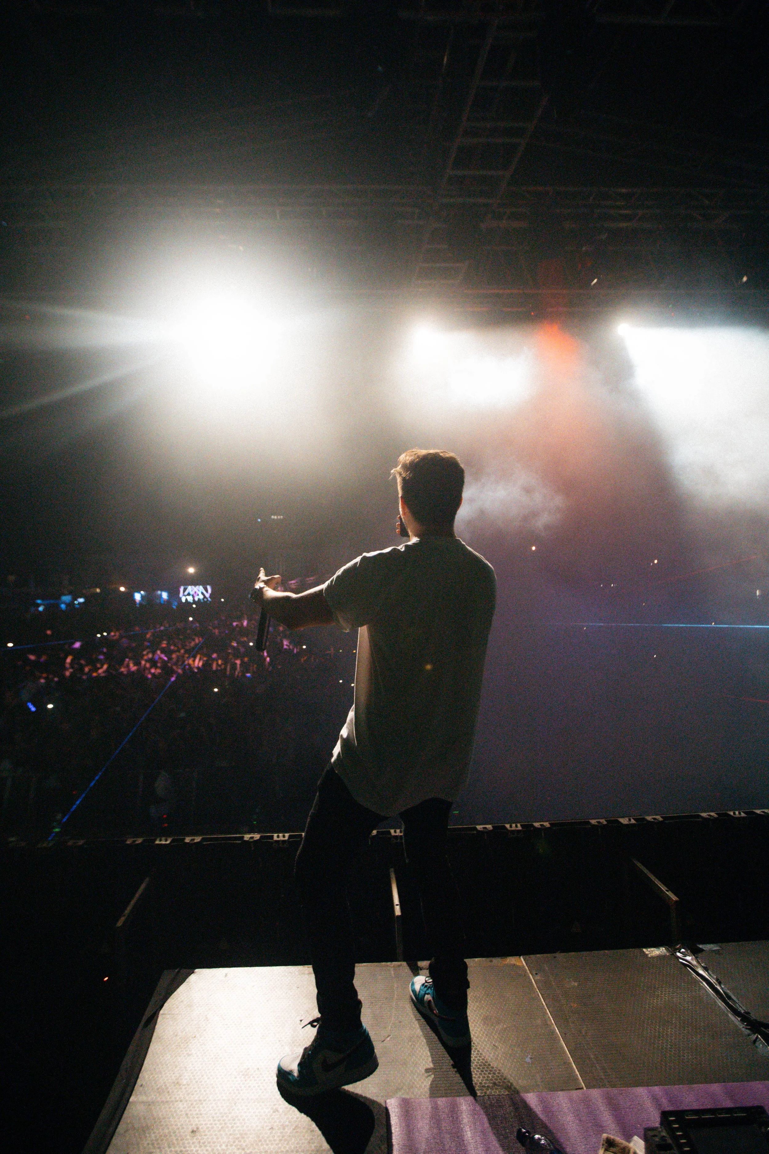 Performer on stage during a concert, viewed from behind, with bright stage lights and an audience in the background.