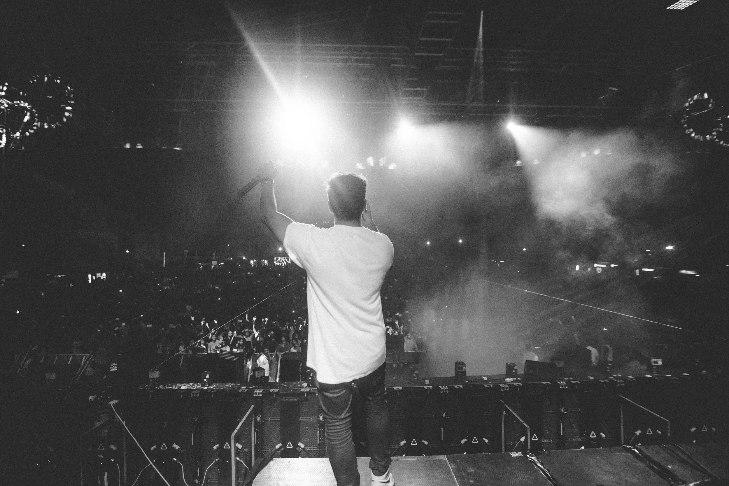 Backstage view of a DJ or performer with short hair, holding a microphone in the right hand, facing a large crowd in a concert or music festival. Bright lights are shining from above, creating a vibrant atmosphere.