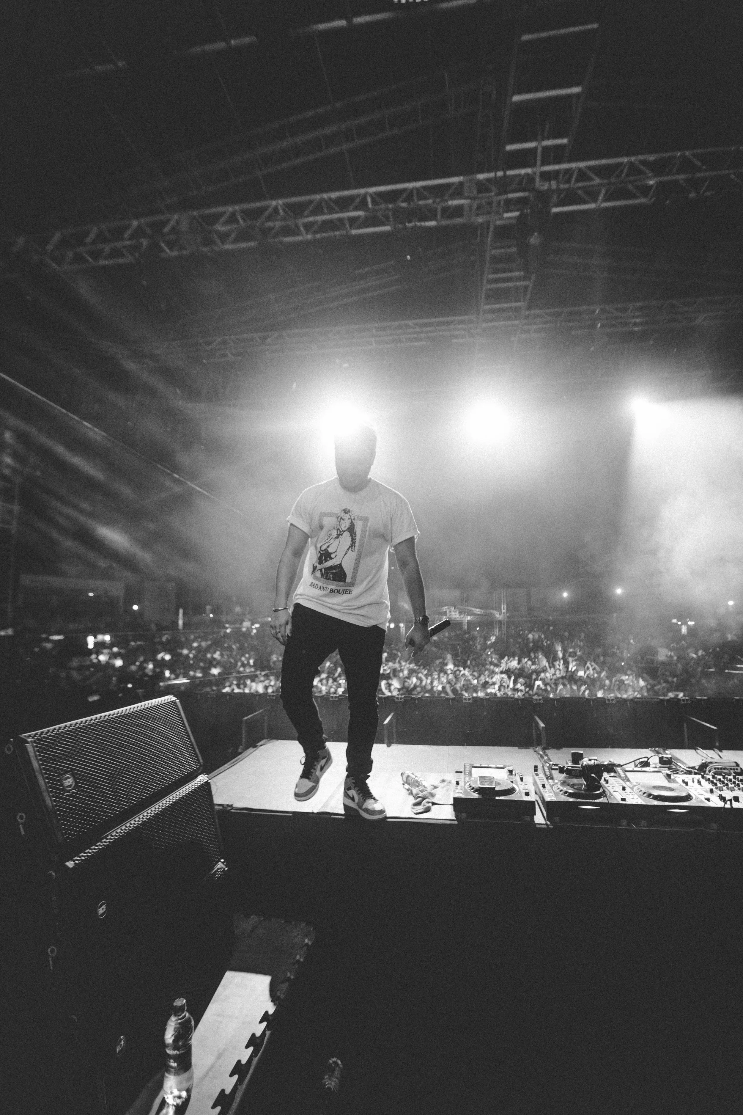 A DJ on stage at night with a large crowd in the background, illuminated by bright stage lights.
