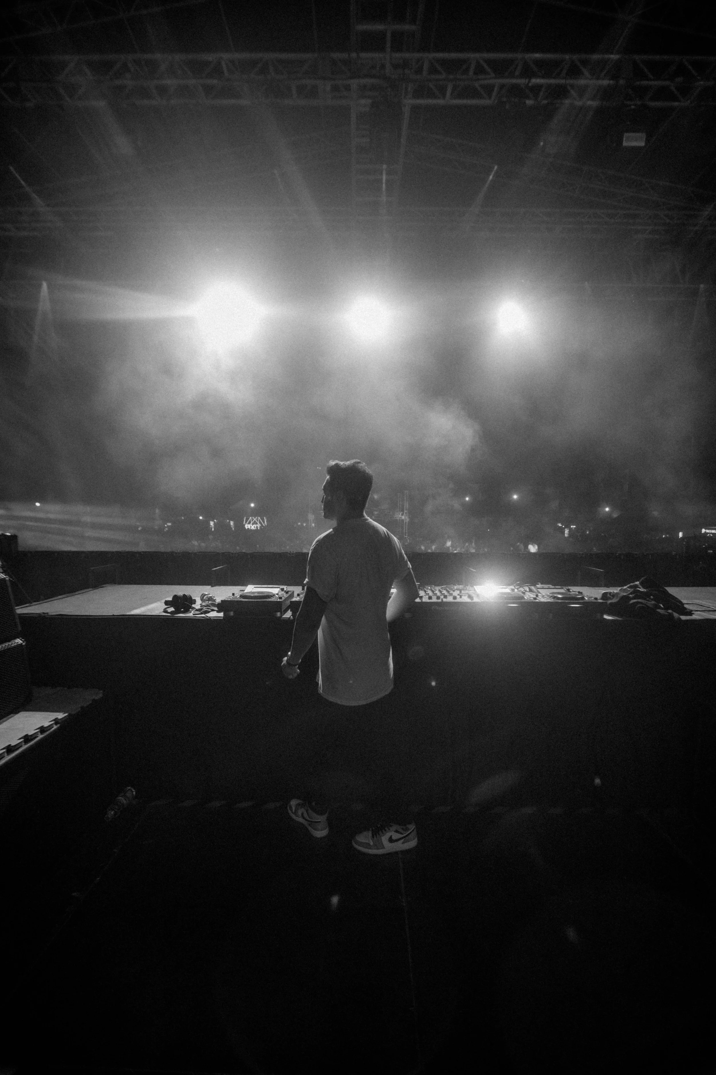 A DJ stands at a mixing console on stage in front of bright stage lights at a concert or music event.