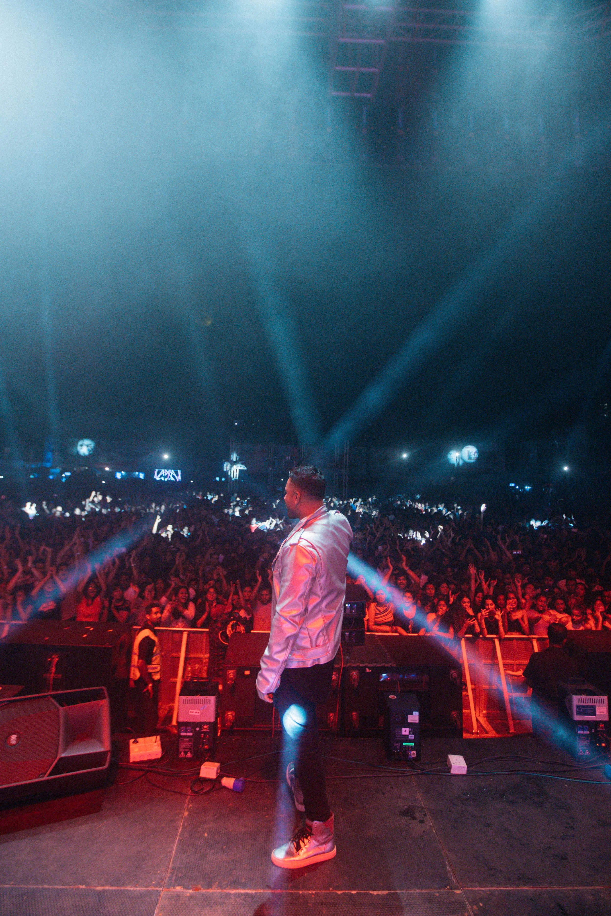 Male performer on stage facing a large crowd at a concert or music event, with colorful stage lighting and a dark backdrop.