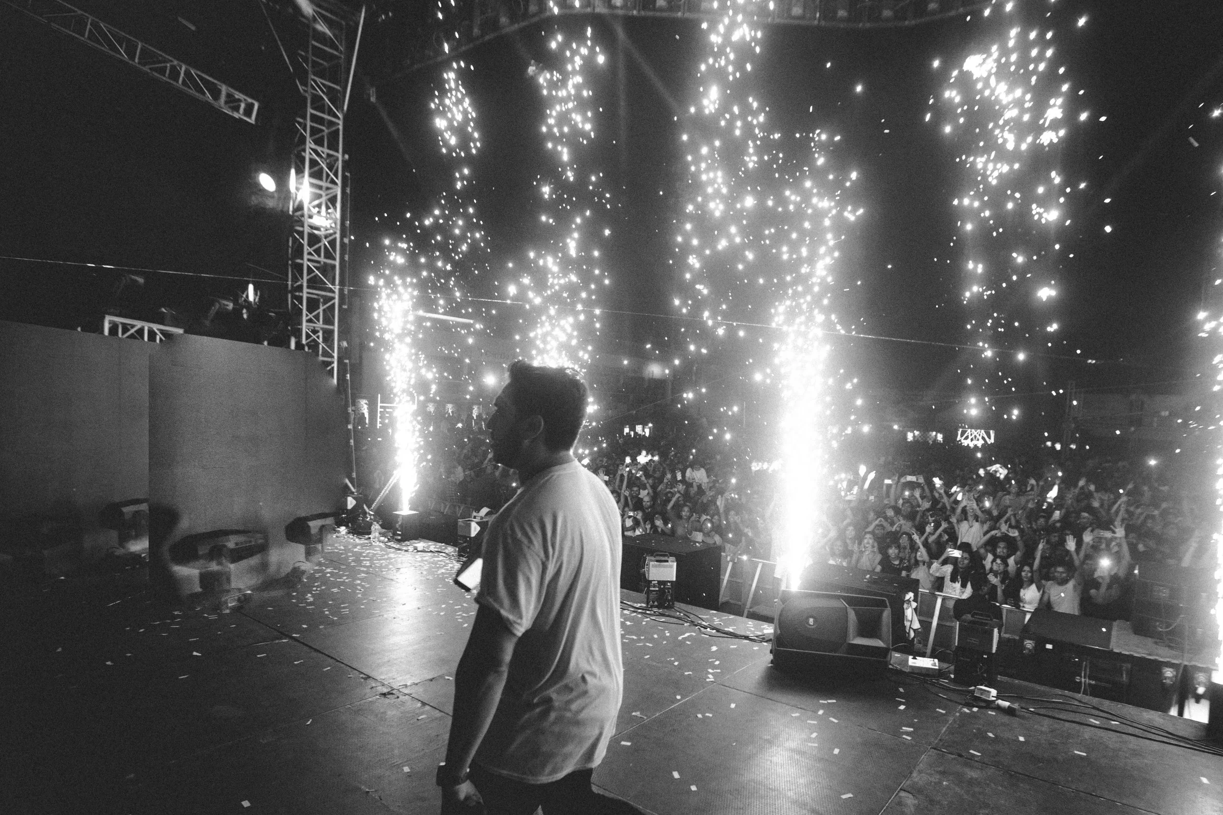 A person standing on a stage at a party or celebration, with fireworks or sparklers in the background and a crowd of people celebrating.