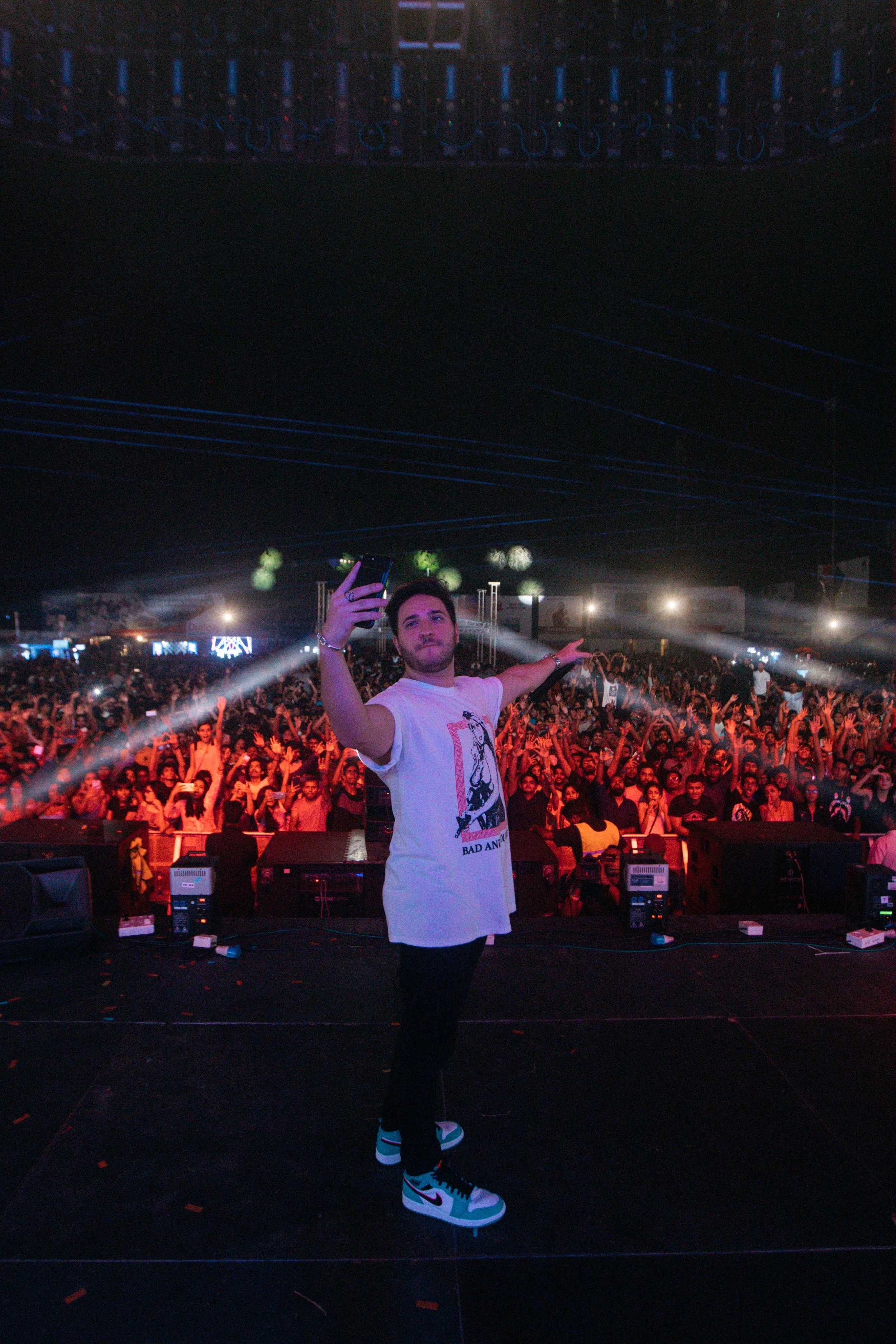 A young man taking a selfie on stage during a concert or festival, with a large crowd of people in the background enjoying the event.