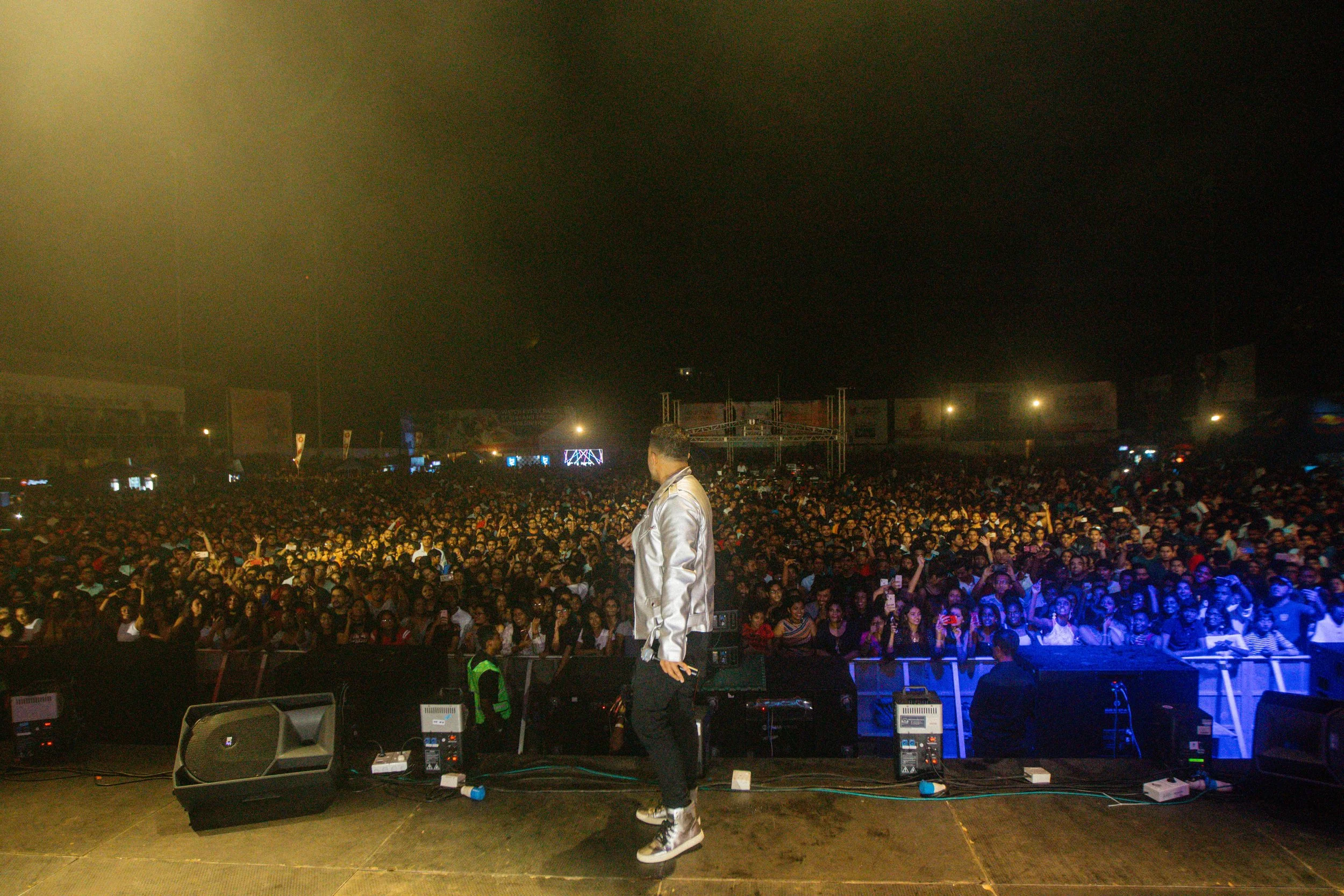 Performer on stage at a concert, facing a large crowd during nighttime with some stage lighting and equipment visible.