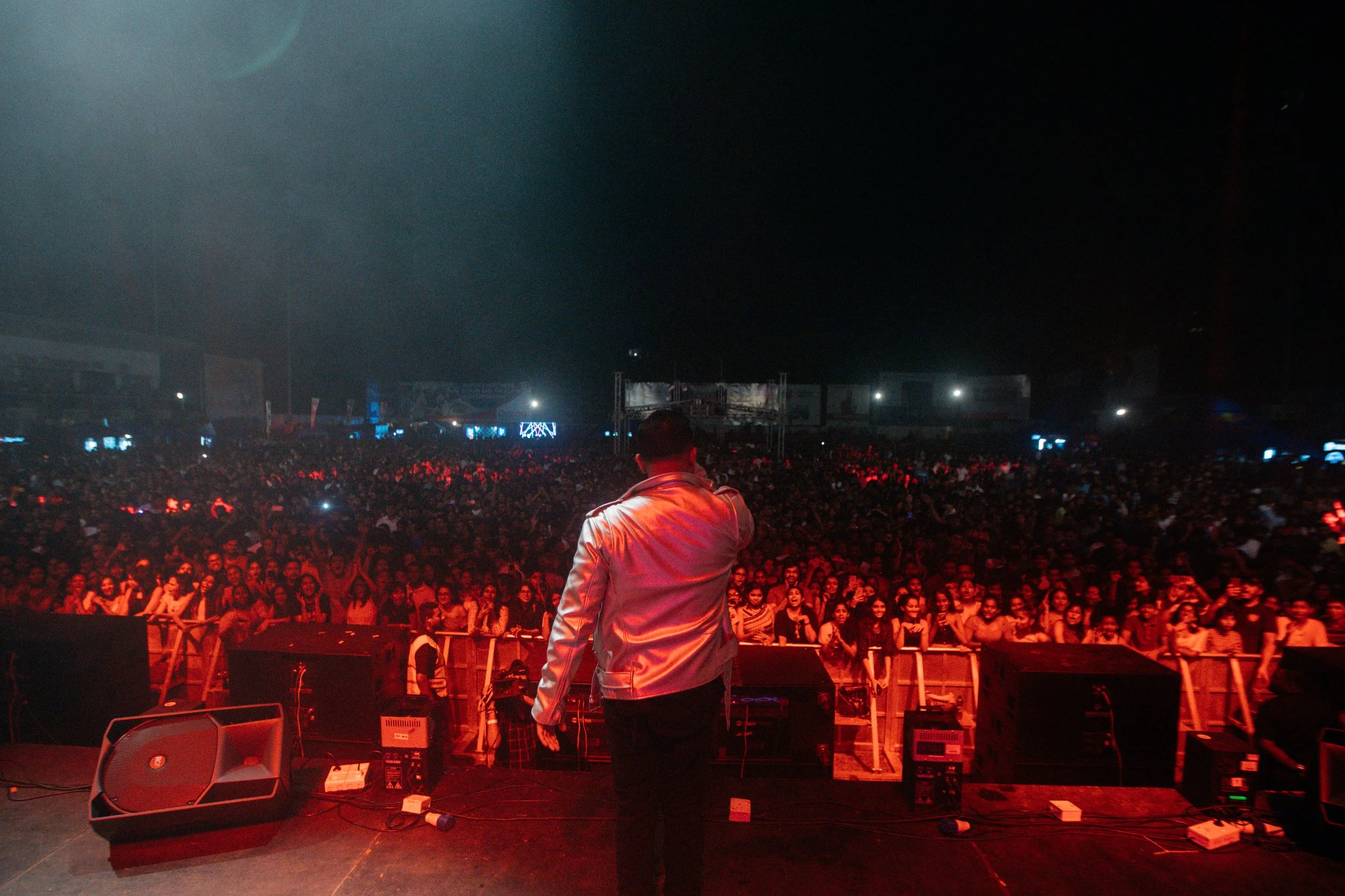 A performer on stage facing a large audience at a concert or event at night, with equipment and speakers in the foreground.